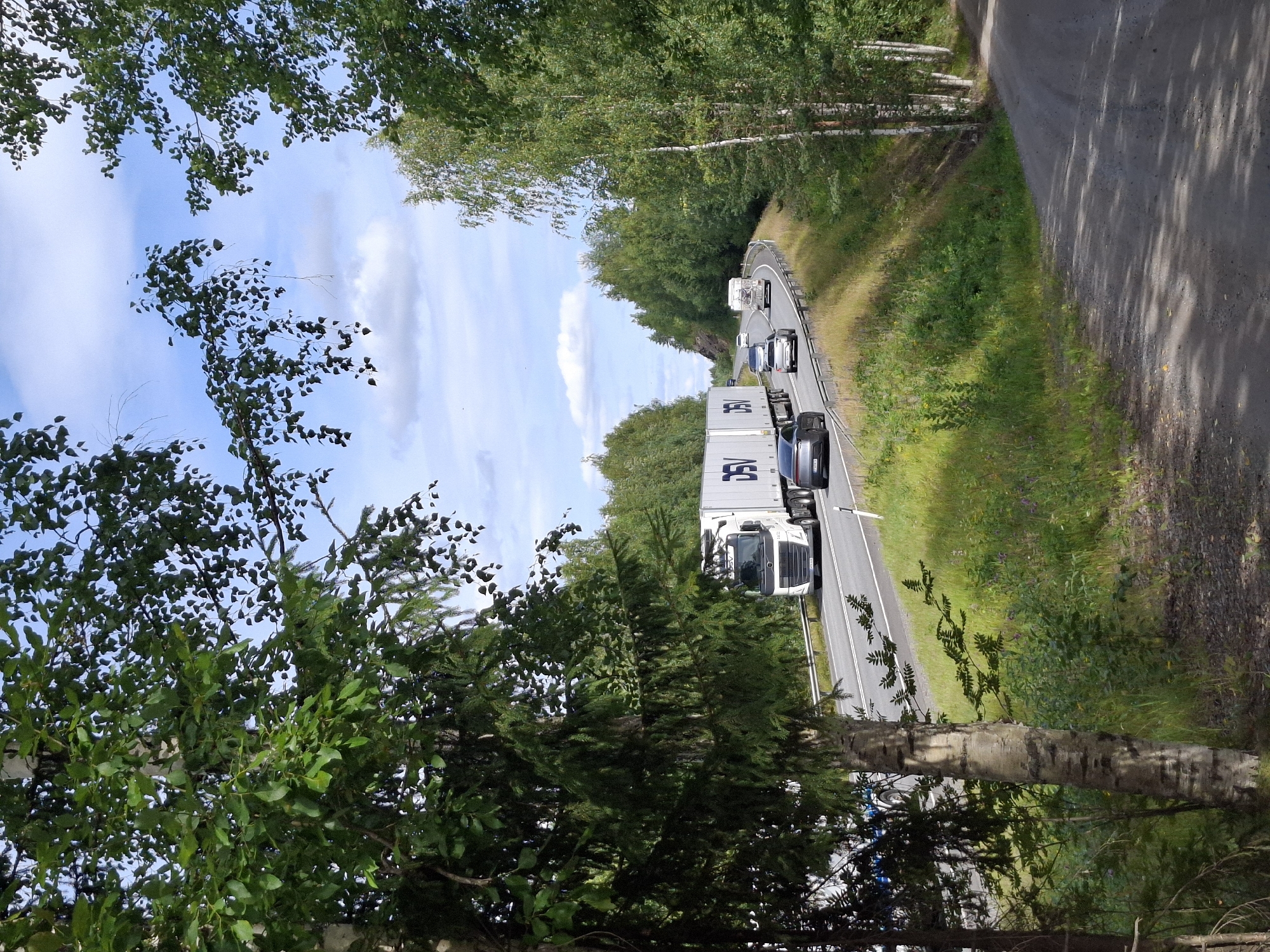 A white semi-trailer truck with "DSV" logo and two white box trailers drives on a highway in Mikkeli, Finland. The truck is in the right lane. A dark gray sedan is in the left lane behind the truck. A silver sedan is in the right lane behind the truck. A white RV is in the right lane in the distance. The highway is surrounded by lush green trees and grass. The sky is blue with scattered white clouds. A dirt road is visible in the foreground to the right.