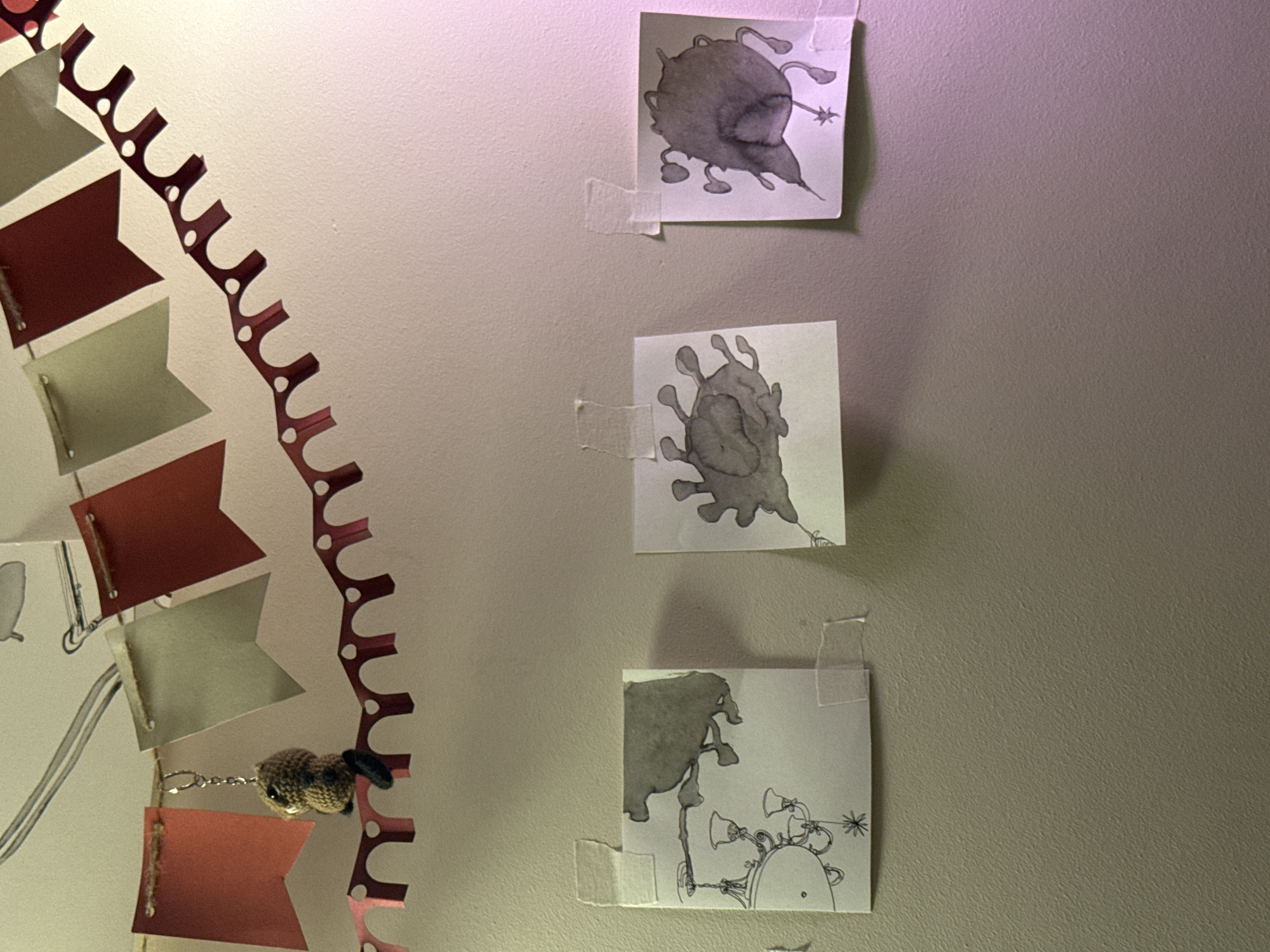 The image shows a section of a wall with three small, square, black and white drawings taped to it. The drawings appear to be abstract or cartoon-like creatures. To the left, a garland of small, triangular flags (alternating red and grey) is partially visible, strung together with twine and adorned with a small crocheted animal hanging from it. The wall itself is a light, solid color. The image does not provide any information regarding the location in Moscow, Russia.