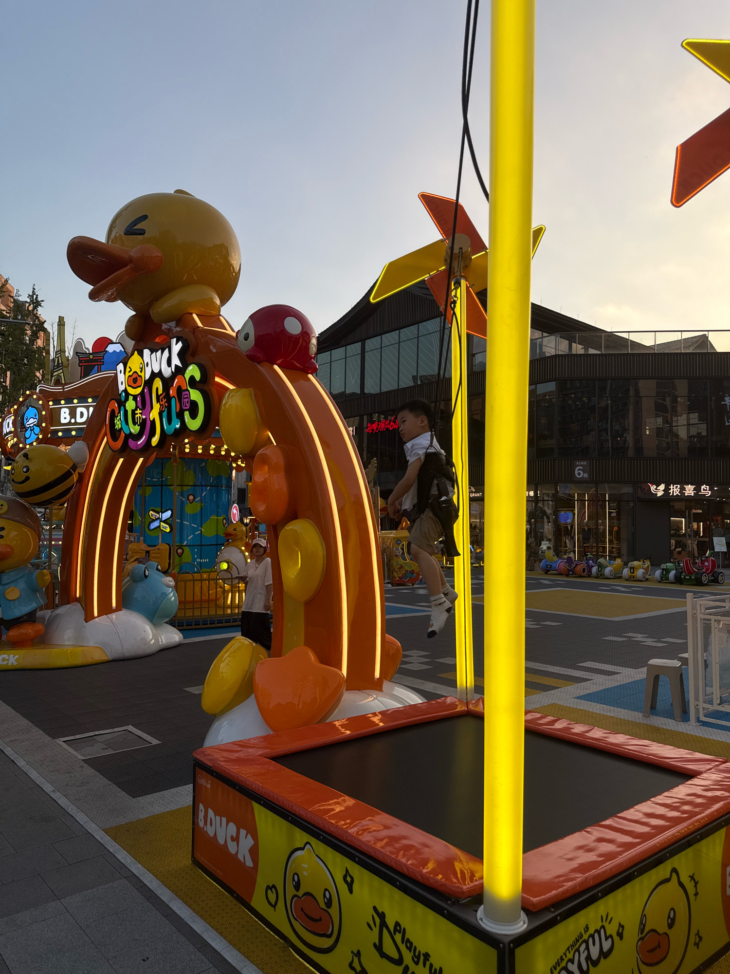 A young boy in a white shirt and shorts is suspended in mid-air, swinging on a rope attached to a yellow pole. The pole is part of a colorful, inflatable-like structure featuring a large yellow duck character and other cartoonish figures, with the text "B.DUCK Cityfuns" prominently displayed. The scene appears to be an outdoor children's play area or amusement attraction, possibly in Yiwu, China, given the signage's stylistic elements and the presence of retail buildings in the background. In the foreground, a trampoline with "B.DUCK" branding is visible. The lighting suggests it is late afternoon or early evening, with the sun casting a warm glow. The background shows modern commercial buildings, some with reflective glass facades, and other smaller amusement rides for children.