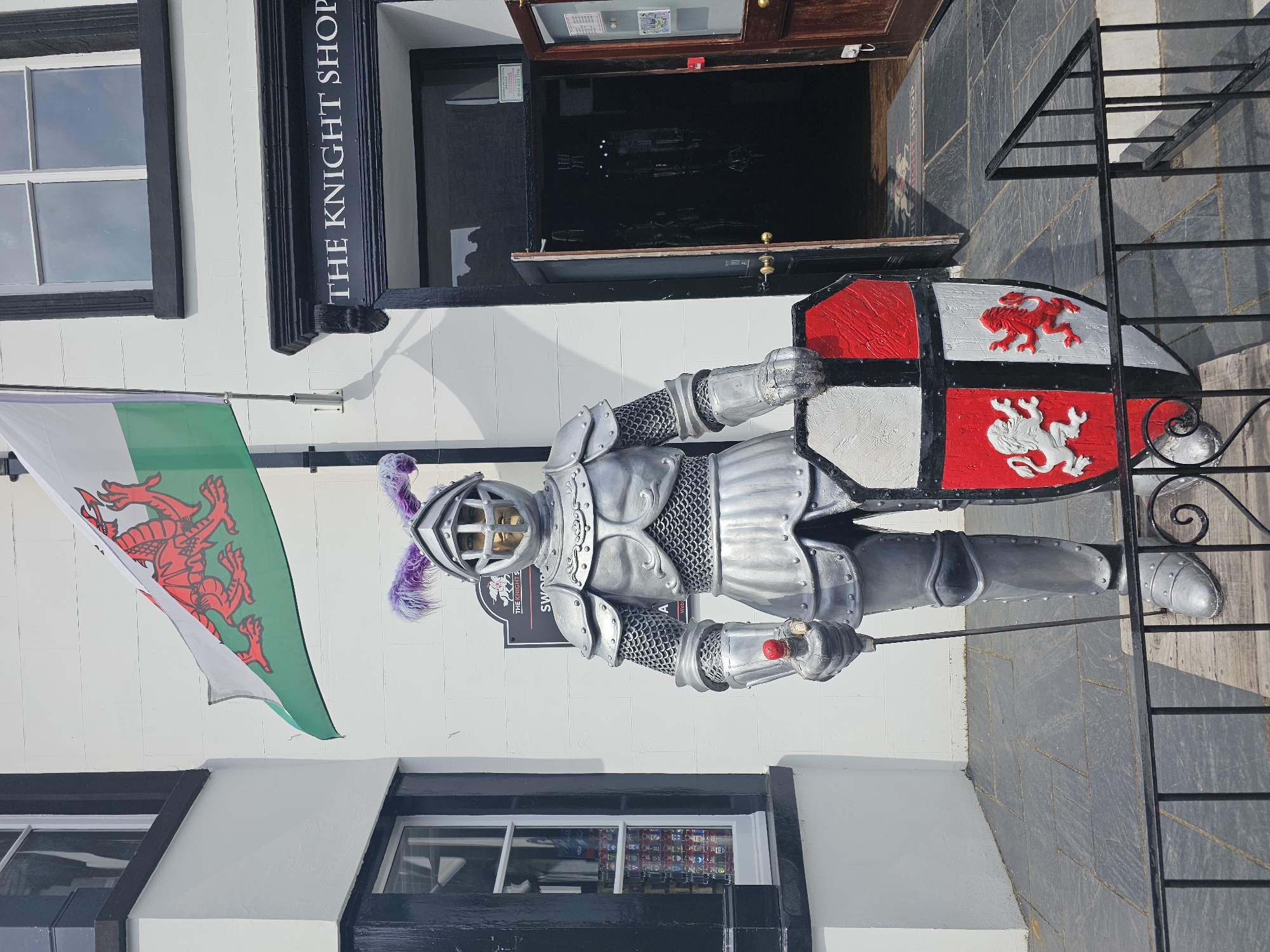 The image depicts a full suit of medieval armor displayed outside a shop named "The Knight Shop." The armor is silver and grey, complete with a helmet, chainmail, breastplate, gauntlets, leg armor, and foot coverings. The knight holds a shield divided into red and white quarters, each with a lion emblem. A Welsh flag hangs to the left of the armor. The shop's black trim and lettering are visible above the armor. The scene appears to be in Llandudno Junction, United Kingdom. The background shows a section of a stone-tiled walkway and a black metal railing. The shop's open doorway reveals a dark interior. Windows are visible on either side of the shop entrance.