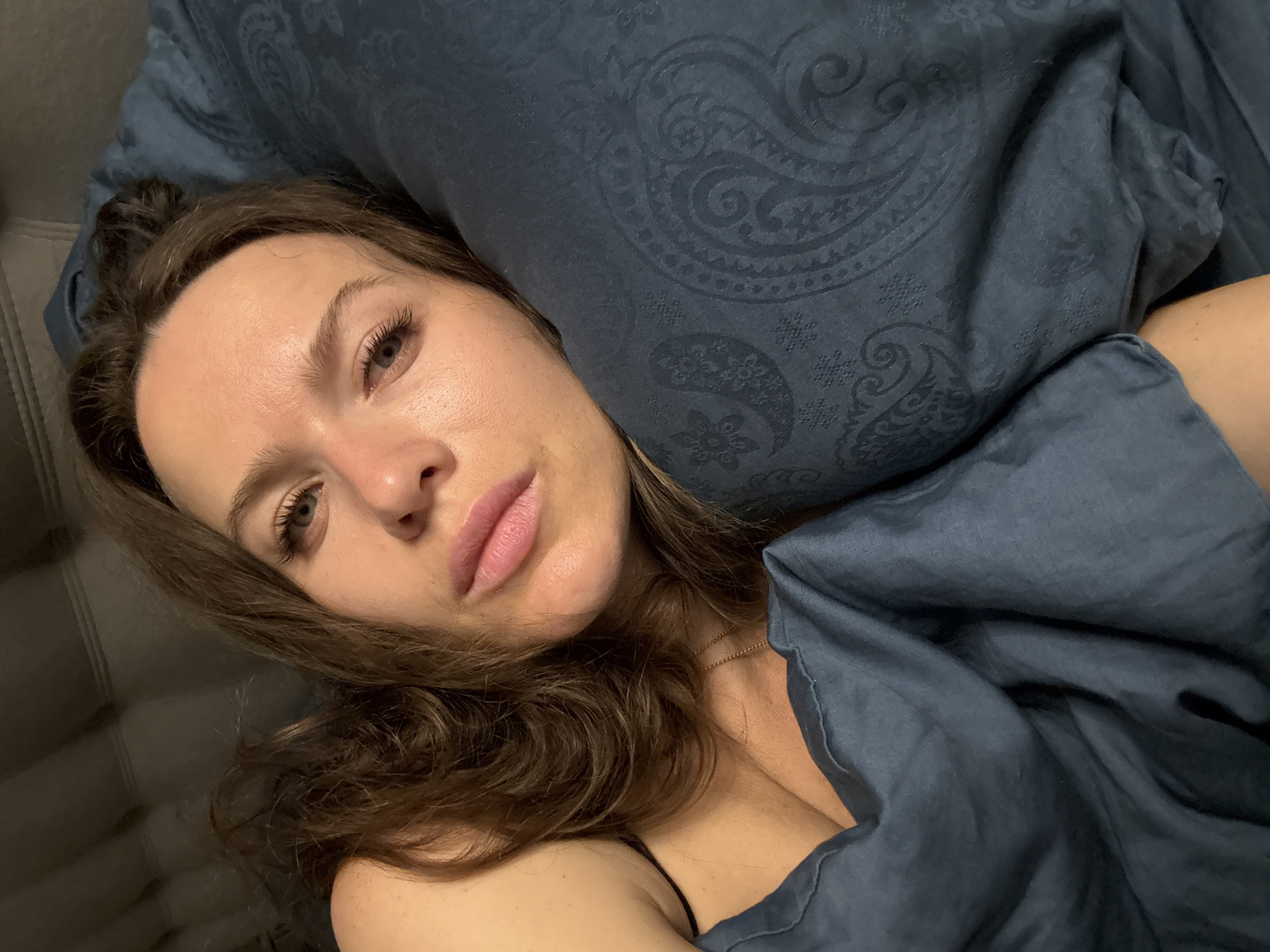 A young woman with light brown hair lies on a grey sofa, resting her head on a blue patterned pillow. A blue blanket is draped over her body. She is looking directly at the camera. A thin gold chain is visible around her neck. The background is out of focus, obscuring details of the location. There is no information to confirm that the image was taken in Khimki, Russia.