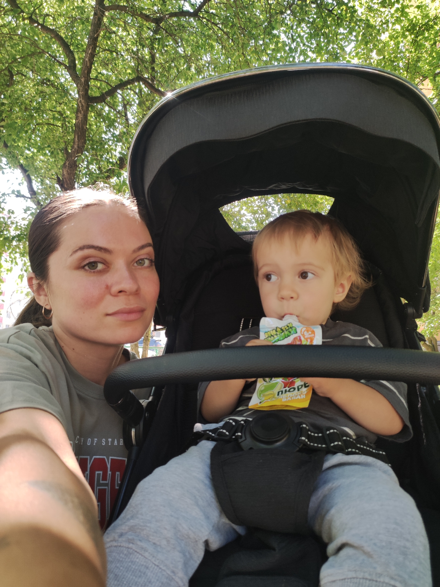 In the city of Chaikovsky, Russia, a woman with dark hair pulled back into a ponytail and green eyes is taking a selfie in a stroller. She is wearing a grey t-shirt. Next to her, a young child with blond hair is seated in the stroller, holding a pouch of juice and drinking from it. The child is wearing a grey and white striped shirt and light grey sweatpants. The stroller has a black canopy and a black safety harness. The background shows lush green trees and dappled sunlight, suggesting an outdoor setting, possibly a park or a tree-lined street.