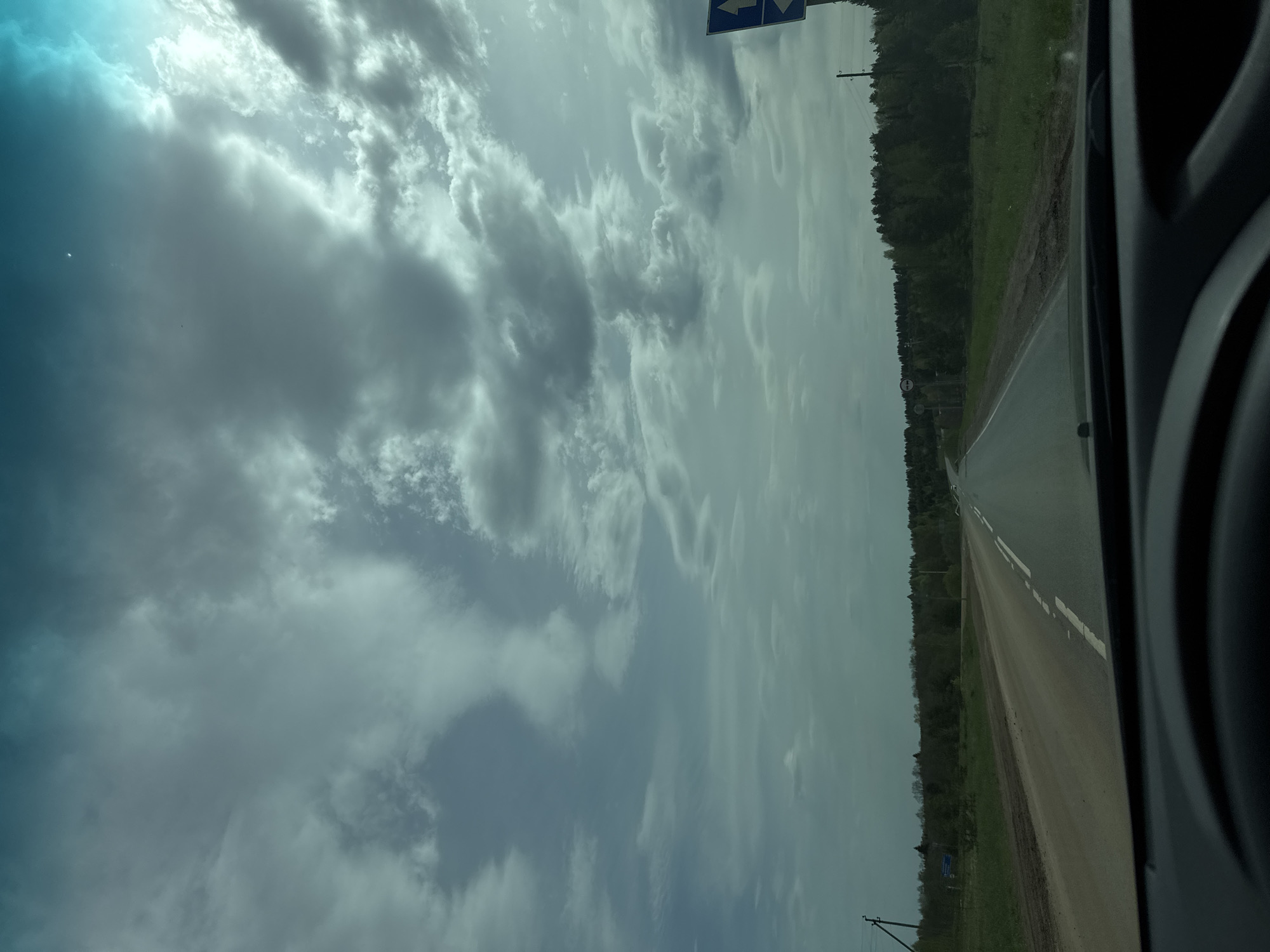 The image shows a view from inside a vehicle, likely a car, looking out onto a road. The road has a white line dividing the lanes and is flanked by green vegetation and a forest. The sky is filled with clouds. The location may be near Kamennyi Kliuch, Russia based on the prompt.