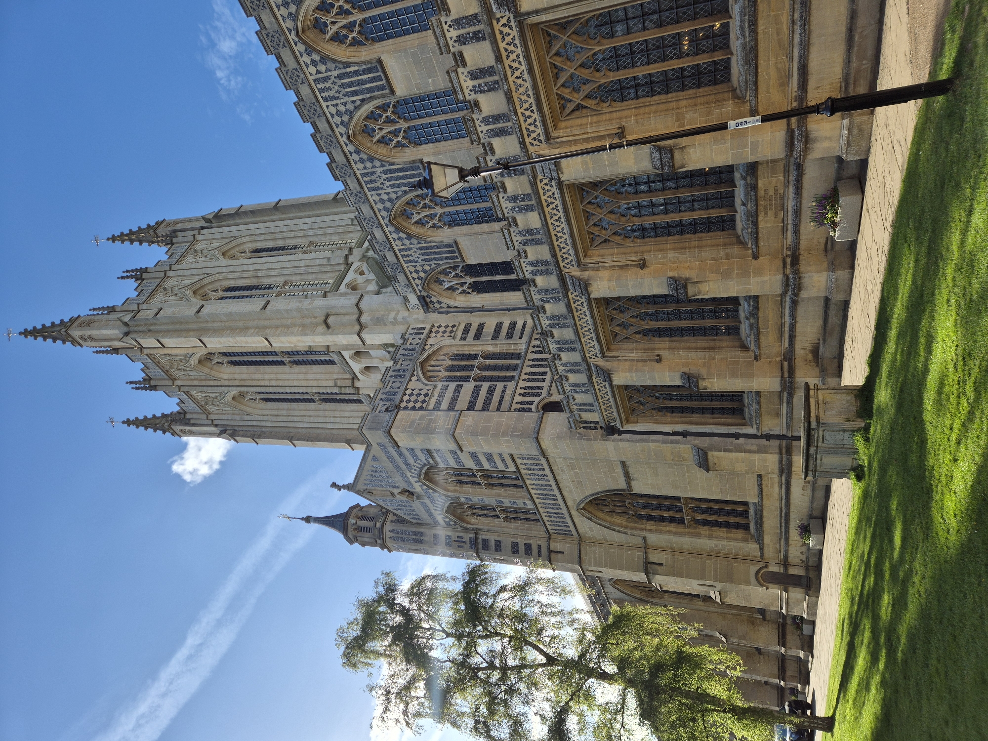 The image shows St Edmundsbury Cathedral in Bury St Edmunds, West Suffolk, United Kingdom. The cathedral's exterior is made of light-colored stone with Gothic architectural details, including pointed arches, ribbed vaults, and intricate window tracery. The building has a tall, ornate tower visible against a bright blue sky with contrails. A street lamp extends from the wall. A tree stands near the base of the building, with green foliage visible. A green lawn stretches out from the building, with a paved walkway along the cathedral wall.