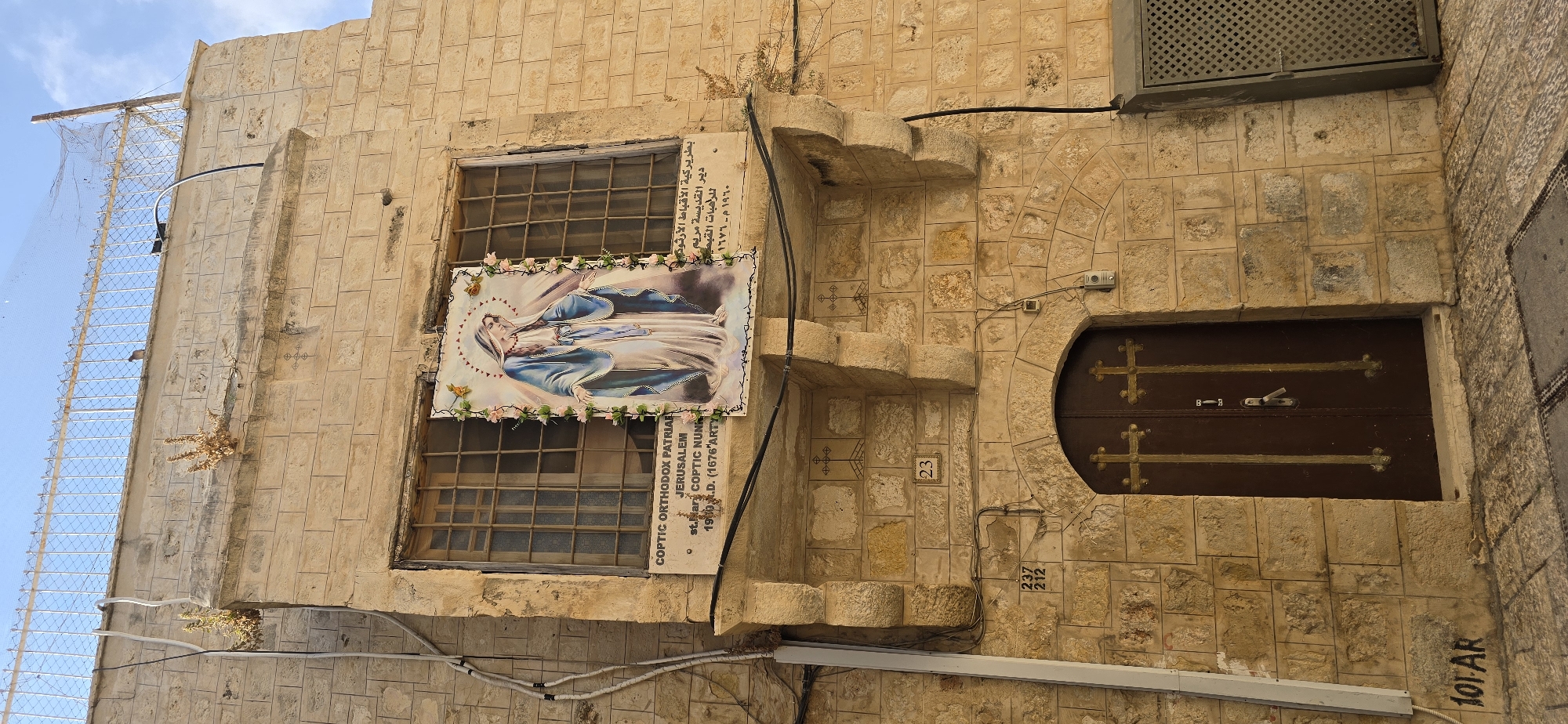 This image captures a close-up view of a section of a stone building in Jerusalem, Israel. The building's facade is constructed from light-colored, roughly cut stone blocks. Above a wooden door with ornate cross embellishments, a sign is affixed to the wall. The sign reads "COPTIC ORTHODOX PATRIARCHATE JERUSALEM st. Mary COPTIC Nunnery 1900 A.D. (1676 Art. D.)". Adjacent to this sign, a large framed picture of the Virgin Mary, depicted in blue and white robes with a halo and surrounded by pink flowers, is hung. The building features a small balcony-like projection with decorative stonework. Above the balcony, a window with barred panes is visible, and on the wall above that, two dried plants sprout from the stonework. A metal fence with a blue sky behind it caps the top of the building. Electrical wires run down the side of the building, and a metal grate is visible to the right of the door. The street in front of the building is paved with stone. The overall scene suggests a historical religious site.