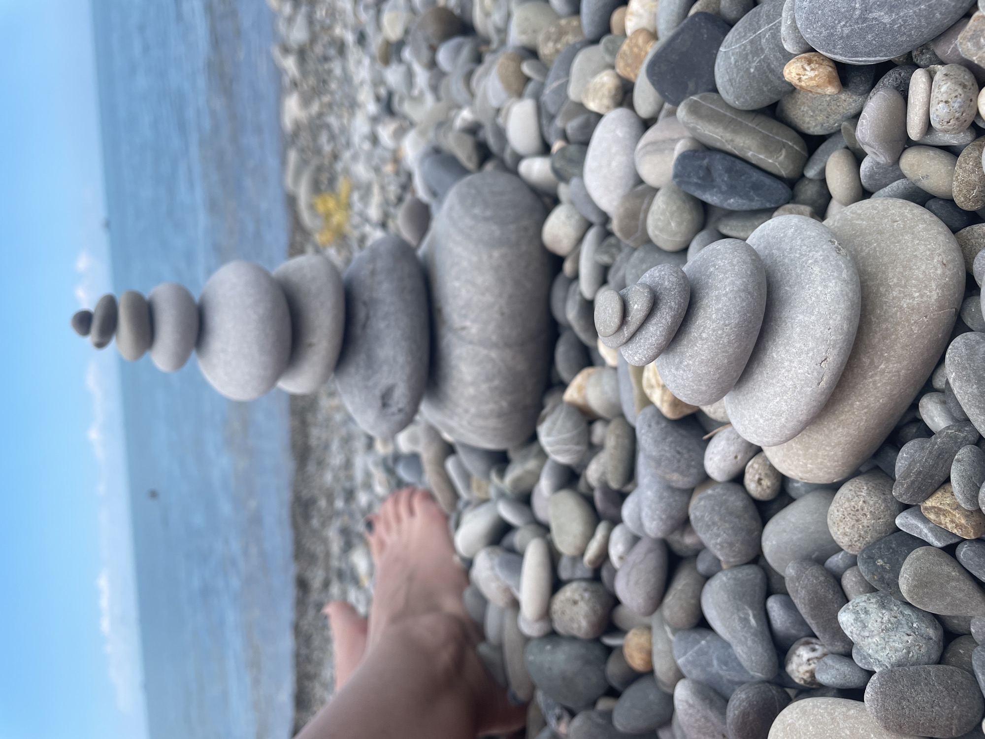 In an outdoor, daylight scene, a person's bare feet are visible on a rocky shore near a body of water. The feet, appearing to be attached to someone sitting just out of frame to the left, are positioned on a surface of small, smooth, gray and tan pebbles. In the mid-ground, a tall, multi-tiered cairn constructed from various sizes of smooth, gray stones is the primary subject, balanced vertically. The stones in the cairn range in color from light gray to darker gray. The background features the calm, blue water of the ocean under a pale blue sky with scattered white clouds. The overall environment is a beach.