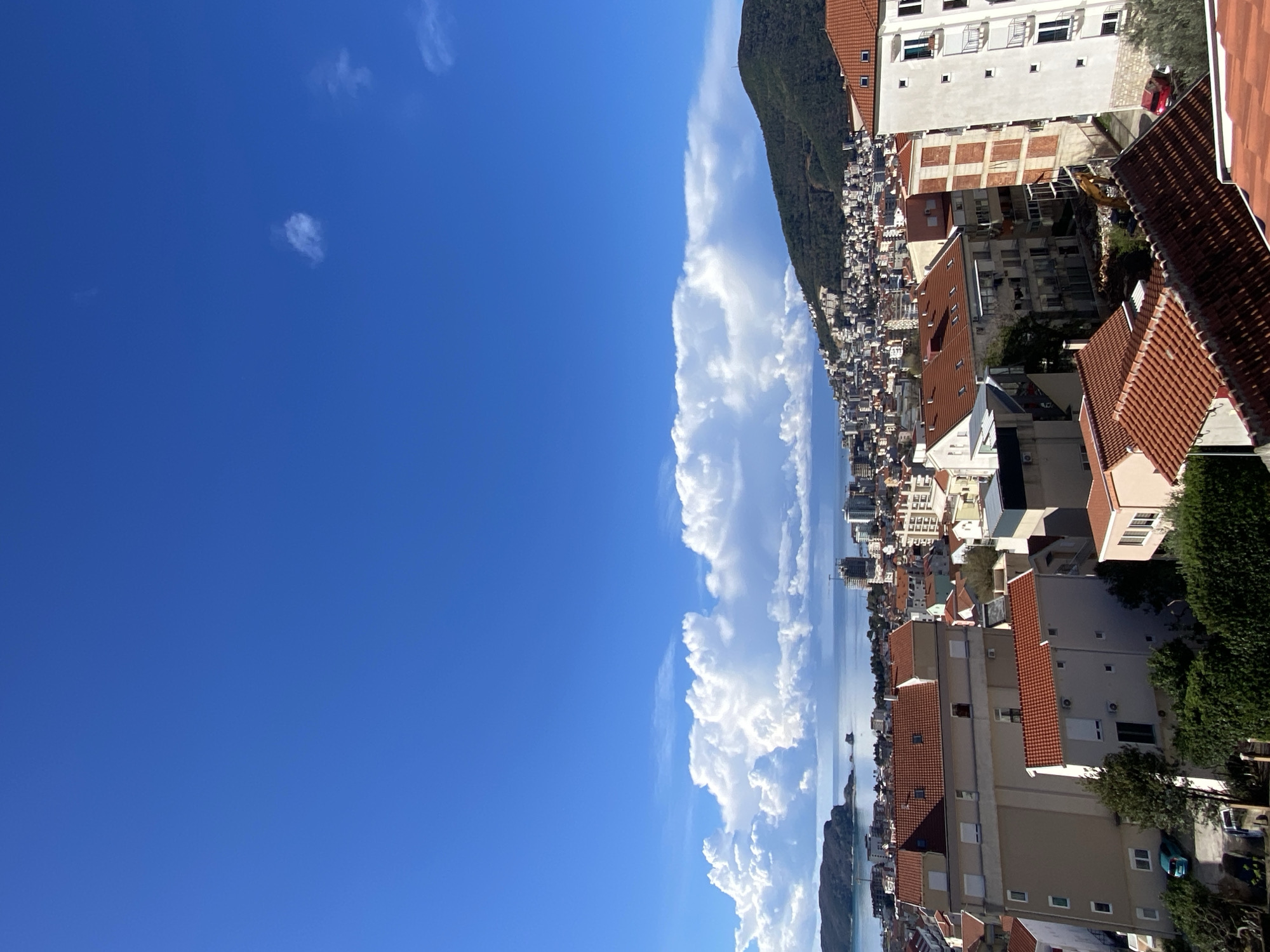 The image displays a panoramic, elevated view of the city of Budva, Montenegro, under a clear, bright blue sky with a prominent band of white cumulus clouds stretching across the horizon above the Adriatic Sea.
In the mid-ground, the city of Budva unfolds, characterized by dense urban development featuring numerous multi-story buildings with predominantly white, beige, and light-colored facades, all topped with terracotta-colored tiled roofs. The city extends along the coastline and climbs the lower slopes of the surrounding green, forested mountains. To the far left, a dark, rocky island or peninsula is visible within the bay. High-rise construction activity, indicated by partially constructed buildings and cranes, is visible along the coast in the distance.
The foreground is dominated by residential structures. On the left, a multi-story building with a light beige facade, white window frames, and a red tiled roof is prominent. A smaller, lighter building with a matching roof sits below it. To the far right, a section of a tall white building with multiple balconies is visible. Between these foreground structures, a localized construction site is identifiable, featuring exposed earth and what appears to be an excavator. Several parked vehicles, including a light blue car on the left and a red car on the right, are visible in small parking areas among the foreground buildings.
No individuals or specific human interactions are discernible within the frame. The scene is static, under bright daylight conditions, suggesting fair weather.