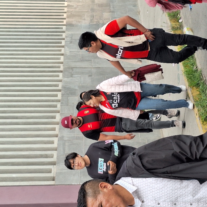 A group of five people stand outside a building. A man wears a red and black striped shirt with a white scarf. Behind him, a woman wears a similar shirt under a white jacket. Another man wears a similar shirt with a red baseball cap. In front of them, a man wears a black Levi's sweatshirt. Finally, another man is wearing a white and gray patterned button up shirt. The building's exterior features a modern, slatted design.