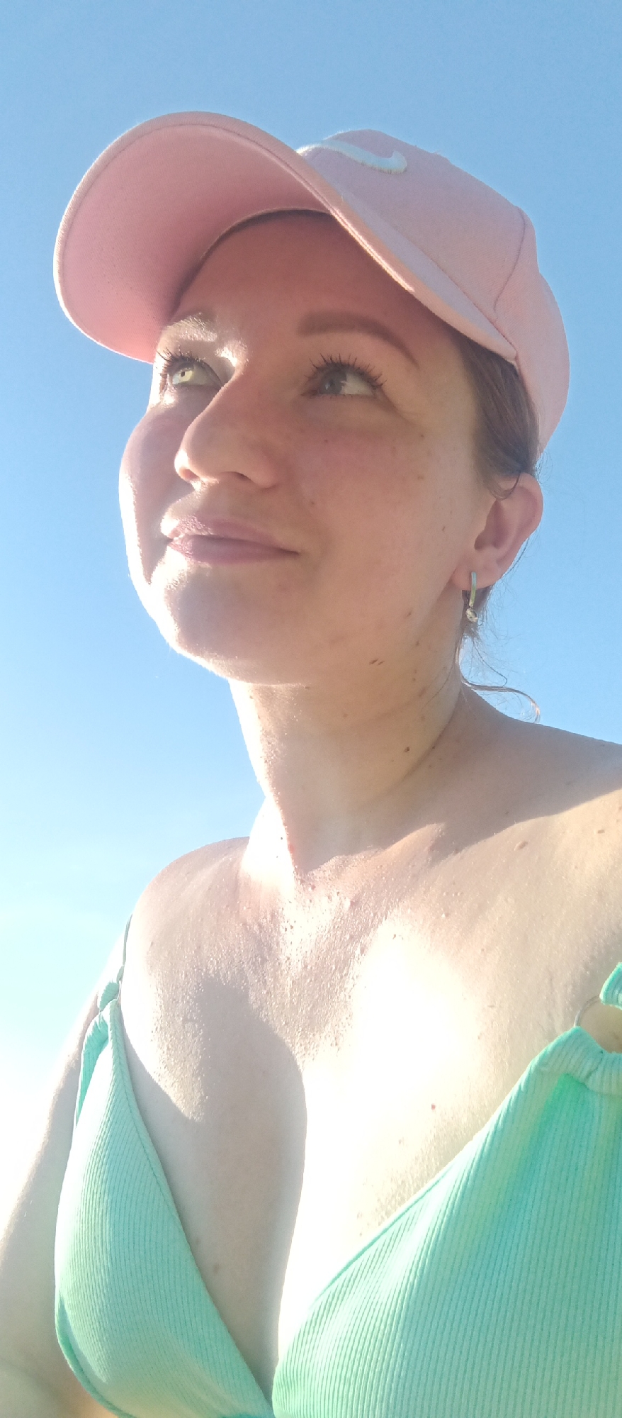 A woman wearing a pink baseball cap and a light green bikini top is gazing upwards in an outdoor setting. The background is a clear, bright blue, suggesting a sunny day. The location appears to be near water, possibly a beach. The city location is unknown.