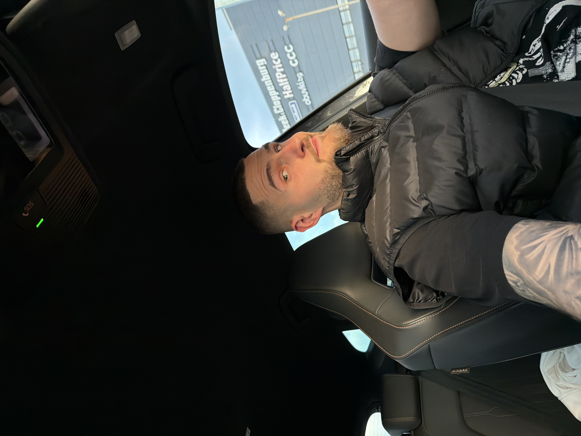 The image shows a man with a trimmed beard and short hair sitting in the passenger seat of a car. He is wearing a black puffer jacket over a black shirt, and a tattoo is visible on his arm. The car appears to be parked in front of a building with the sign "CCC HalfPrice" and "obuvki.bg", indicating a shoe store. The text "eek. Cloppenburg" is also visible on the sign. Based on this, the location is likely in Sofia, Bulgaria, as "obuvki.bg" is a Bulgarian online shoe retailer and CCC is a shoe store chain with locations there. The car interior is black with a leather seat featuring orange stitching. An "AIRBAG" label is visible on the side of the seat.