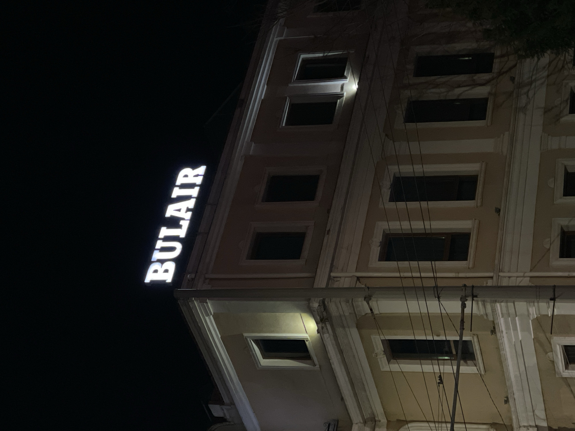 At night, a building is visible, likely in Burgas, Bulgaria based on the "BULAIR" sign. The building is several stories high with rectangular windows, most of which are dark. The building is lit in some areas, highlighting architectural details like molding. Power lines are visible in the foreground. The word "BULAIR" is illuminated in large, vertical, white lettering on the left side of the building. The background is dark, indicating a night scene.