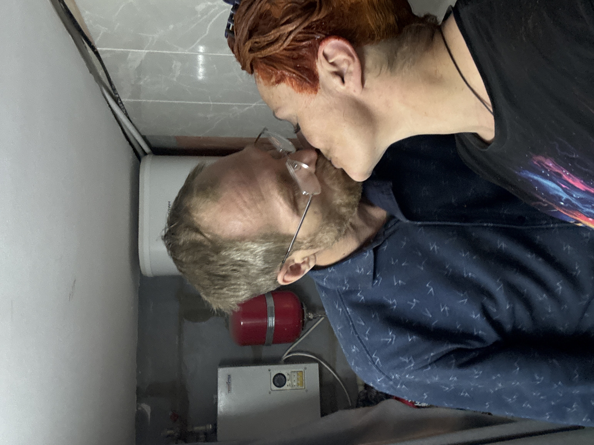 Two people are in a small, enclosed space with plumbing and electrical equipment. One person with red-orange hair is standing and kissing the other person with blond hair and glasses. The location is in the city of Chaikovsky, Russia. The background includes a white water heater, a red expansion tank, and a grey electrical box. The person kissing is wearing a black t-shirt. The other person is wearing a dark blue shirt with a light-colored pattern. The walls are covered in grey tiles.