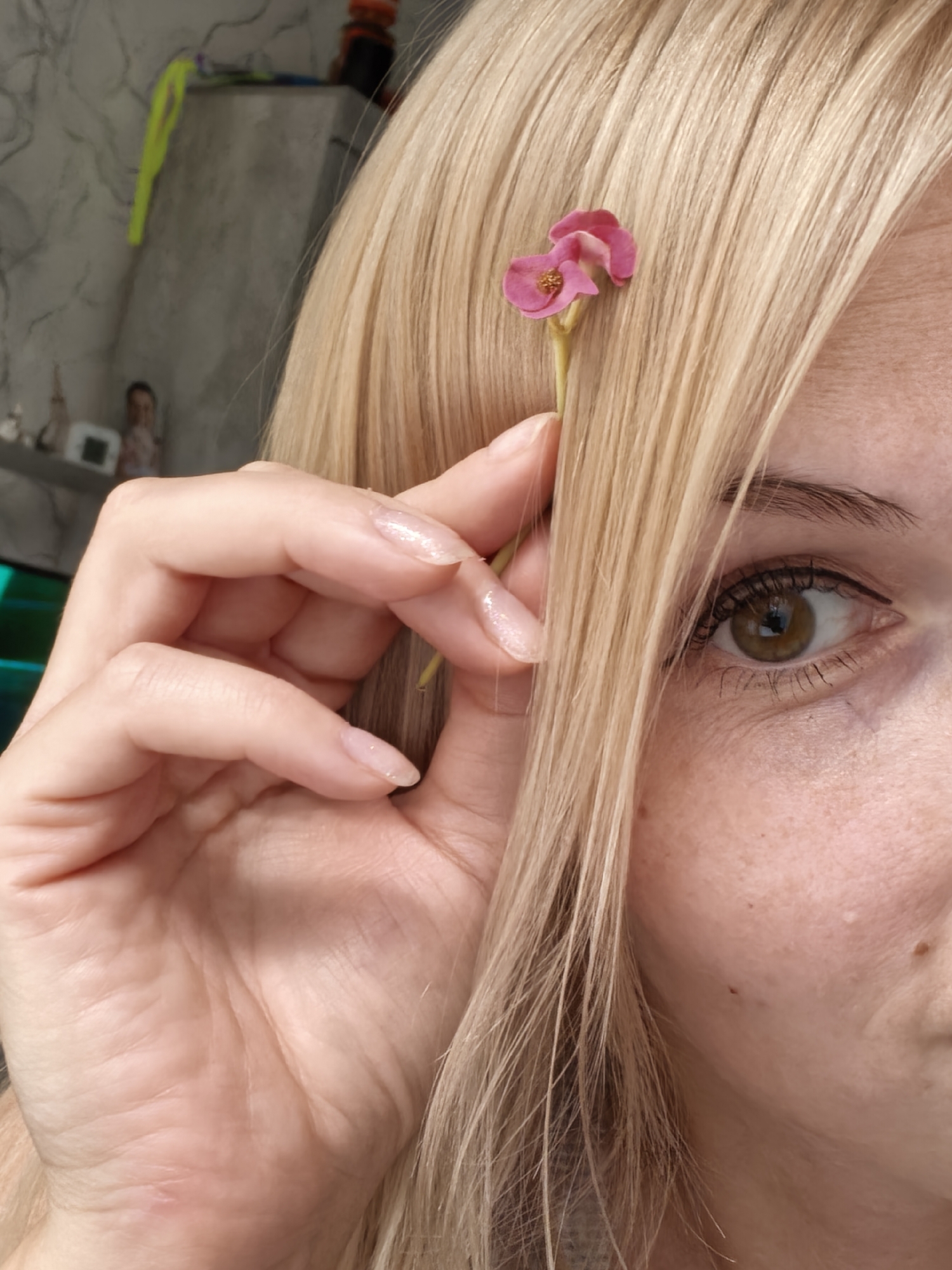 A close-up, eye-level, medium shot captures the right side of a Caucasian woman's face and her left hand, against a blurred background. She has blonde hair, brown eyes, and fair skin with freckles. A small pink flower is held near her hair by her left hand, which has clear, glittery nail polish. The background shows a closet with various items, suggesting an indoor setting. There is no information or visual cues to indicate the location is in Sterlitamak, Russia.