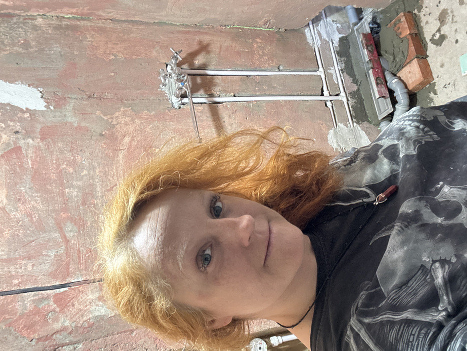 An adult female with bright reddish-orange hair and blue eyes is captured in a selfie in a room undergoing renovation in Chaikovsky, Russia. She wears a dark t-shirt with a grey skull pattern and a black necklace featuring a red, faceted stone pendant. Her expression is a slight smile, and she looks directly at the camera. The background consists of unfinished, textured reddish-brown walls with visible patches of white plaster. On the right side of the frame, a chrome-colored shower mixer rough-in is installed on the wall, with white PVC pipes running vertically and horizontally beneath it. A grey linear shower drain is set into fresh mortar or concrete at the bottom right, with a red spirit level resting on it. Two reddish-brown bricks are partially visible beneath the drain, embedded in the construction material. A white pipe and a partial view of a water meter are visible on the left side of the frame, near the woman's shoulder.