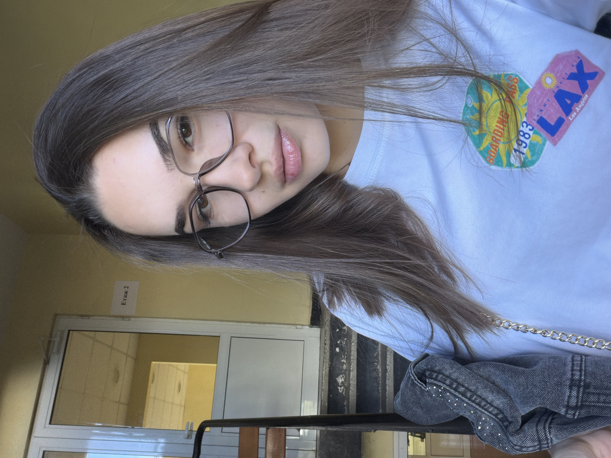 A young woman with long brown hair and glasses is posing for a selfie indoors. She is wearing a white t-shirt with a graphic print, and a denim jacket draped over her shoulder. The background suggests she is in a building with a window or door, a sign in Cyrillic that says "Этаж 2" (Floor 2), and stairs. The location cannot be determined to be in Sofia, Bulgaria from the provided image.