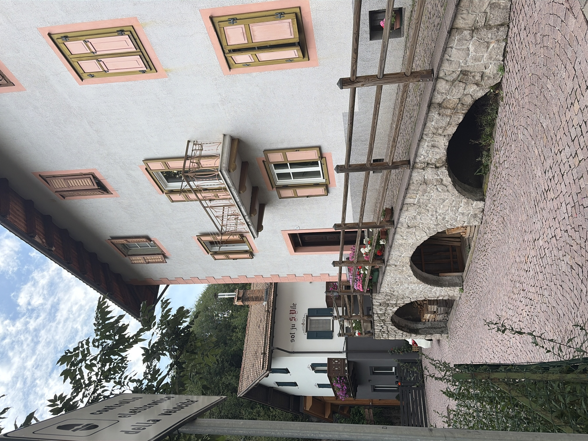 The image depicts a cobblestone street with a stone bridge arching over a lower area. The bridge is lined with a wooden fence and has potted flowers on top. On the right, a pale gray building with pink trim and windows with light pink shutters is visible. To the left, a white building with dark shutters and a sign that reads "sol ju S.Vile" is partially visible. A traffic sign with Italian text is in the foreground. The scene appears to be in Moena, Italy, given the presence of Italian text on the traffic sign and the architectural style consistent with the region. The environment suggests a tranquil, possibly rural or small-town setting, with lush greenery in the background. There are no people or animals visible in the frame.