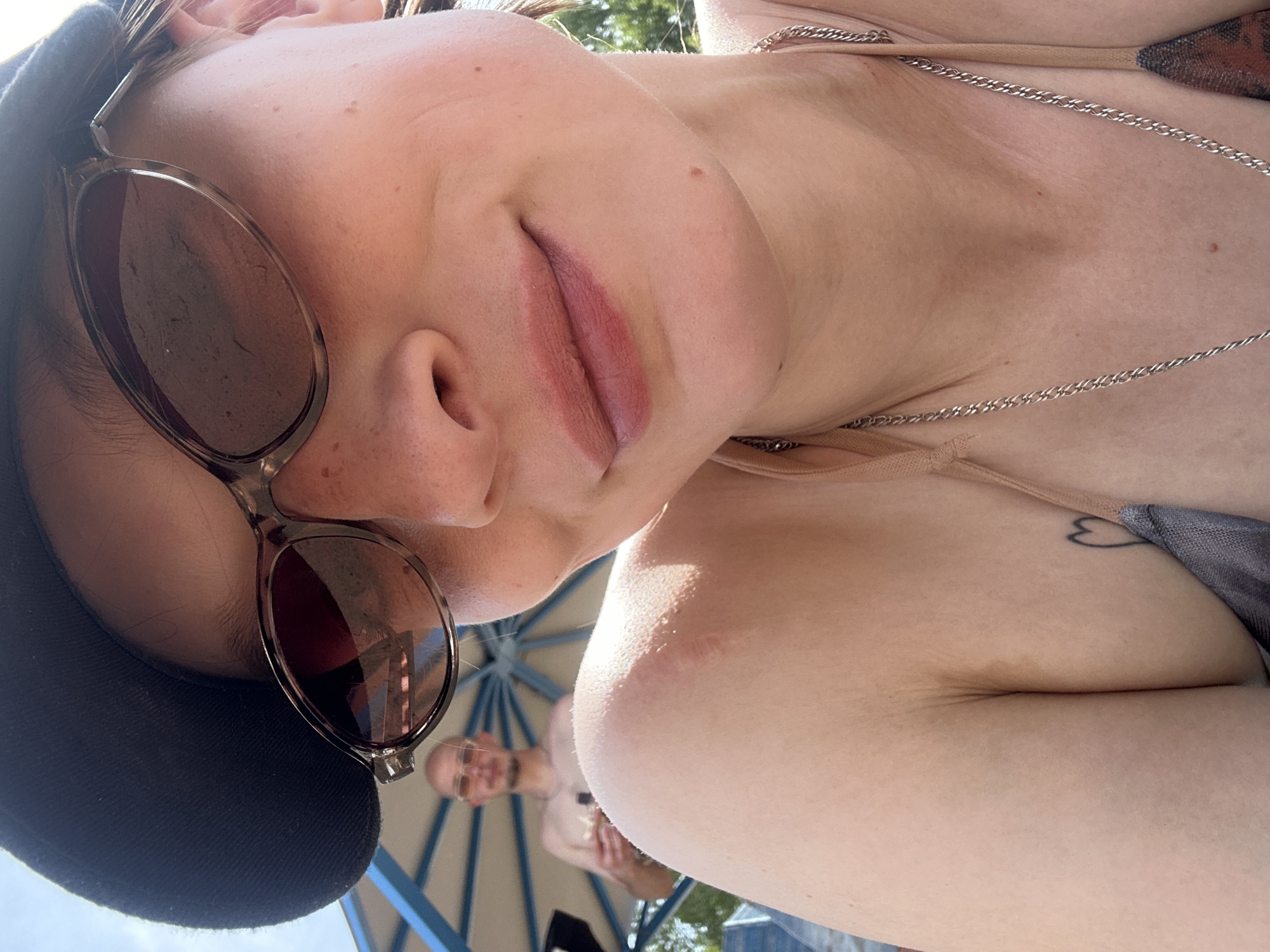 The image shows a close-up of a young woman wearing sunglasses and a black baseball cap, looking at the camera. She is wearing a bikini top. In the background, a man wearing sunglasses and a chain necklace is visible, standing under a blue and white striped umbrella. The setting appears to be outdoors, possibly a beach or poolside area, with greenery visible in the background. The image does not contain any specific geographical indicators that would place it in Chaikovsky, Russia.