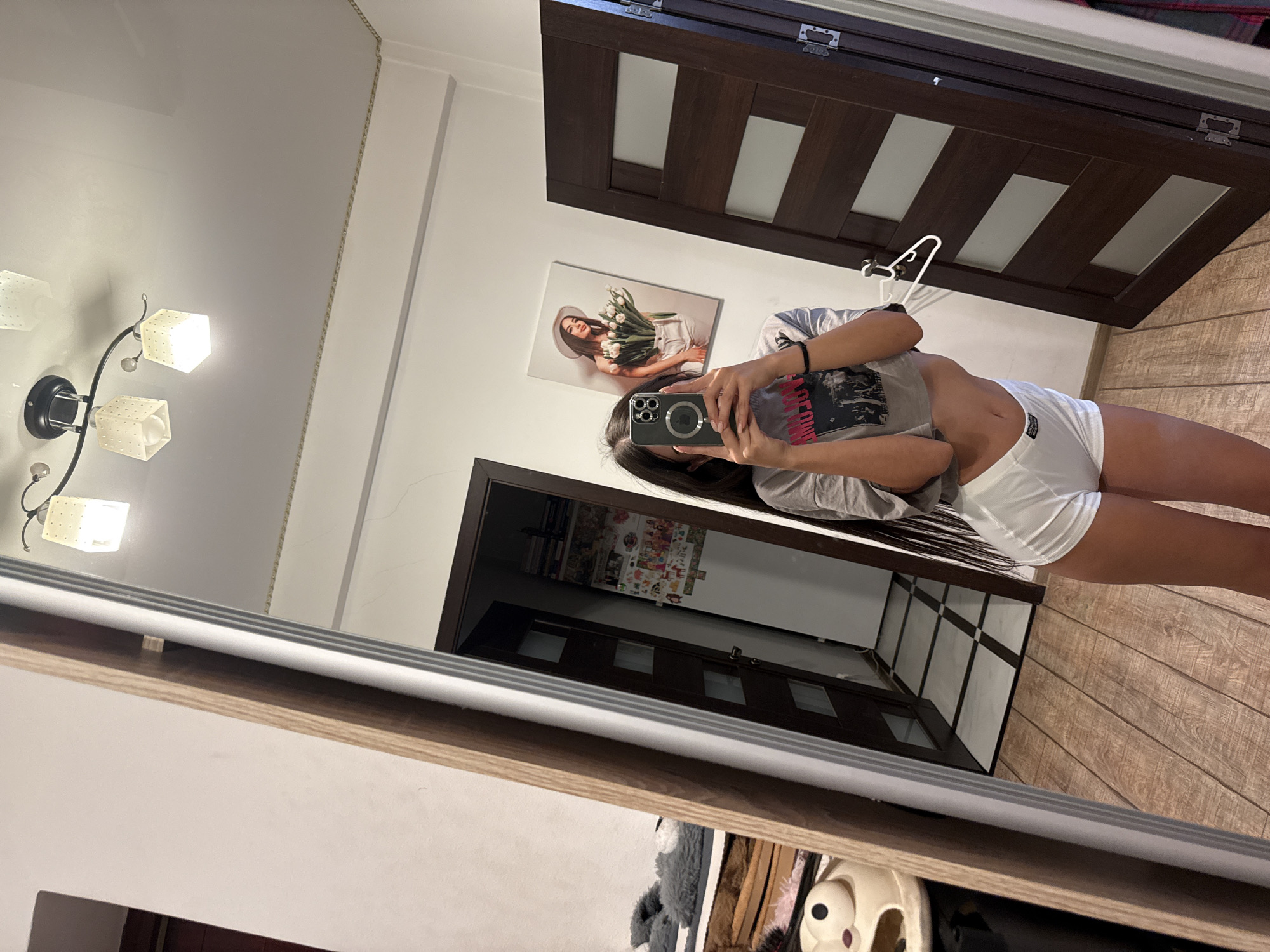 A young woman with long dark hair is taking a mirror selfie in a room. She is wearing a grey crop top with a graphic print and white shorts. She is holding a smartphone in her right hand, angled to capture her reflection. The room appears to be a bedroom, with a wooden floor, a large mirror, two dark wooden doors, and a framed picture of a woman holding tulips hanging on the wall. The reflection also shows a ceiling light fixture with three cube-shaped lamps. In the reflection of the mirror, to the left of the woman, a glimpse of a bed with grey furry slippers and some other items on it can be seen. A doorway in the reflection reveals a hallway with checkered marble flooring. The overall impression is that of a personal space within a residential building. The scene is consistent with an interior residential setting in Chișinău, Moldova, a city known for its diverse architecture, including many apartment buildings with similar interior designs.