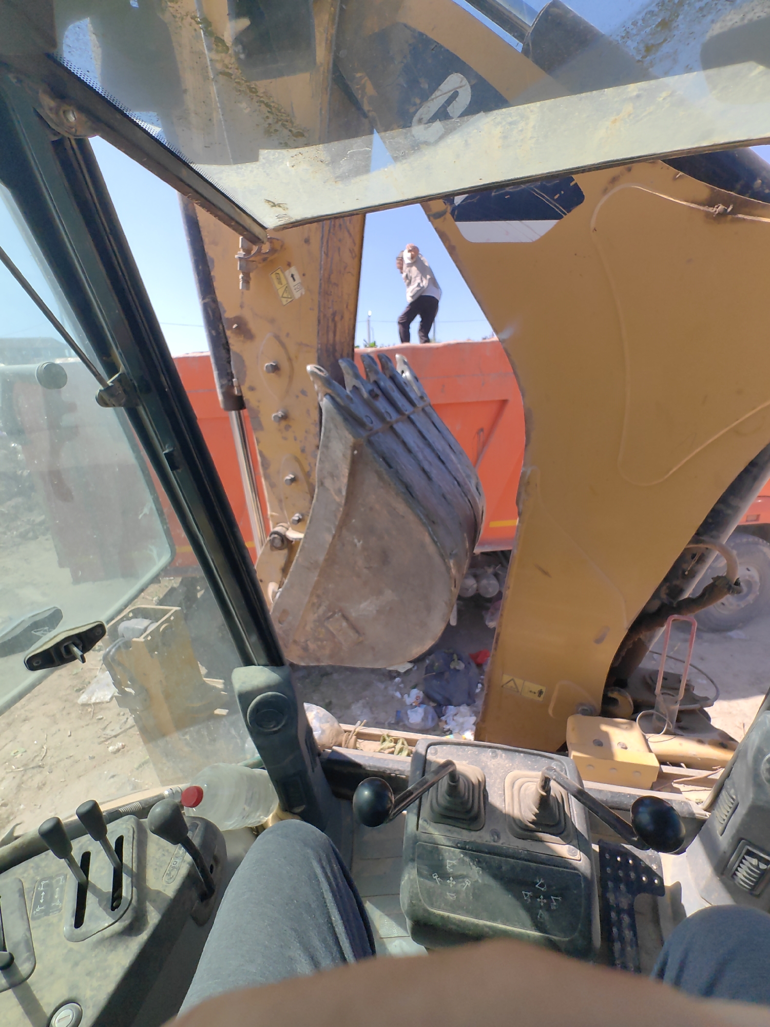 An outdoor, ground-level, first-person perspective shot from inside a construction vehicle, likely an excavator, looking out. The viewpoint is from the driver's seat, with the controls and part of the driver's legs visible in the foreground. The large, metal bucket of the excavator is prominently positioned in the center of the frame, hanging over debris on the ground. In the background, a bright orange dump truck is visible, and a person in a light-colored hoodie and dark pants stands on top of it, looking away from the camera. The environment appears to be a construction or demolition site, with dusty ground, scattered debris, and construction equipment. The sky is clear and blue. The location is specified as Shymkent, Kazakhstan.