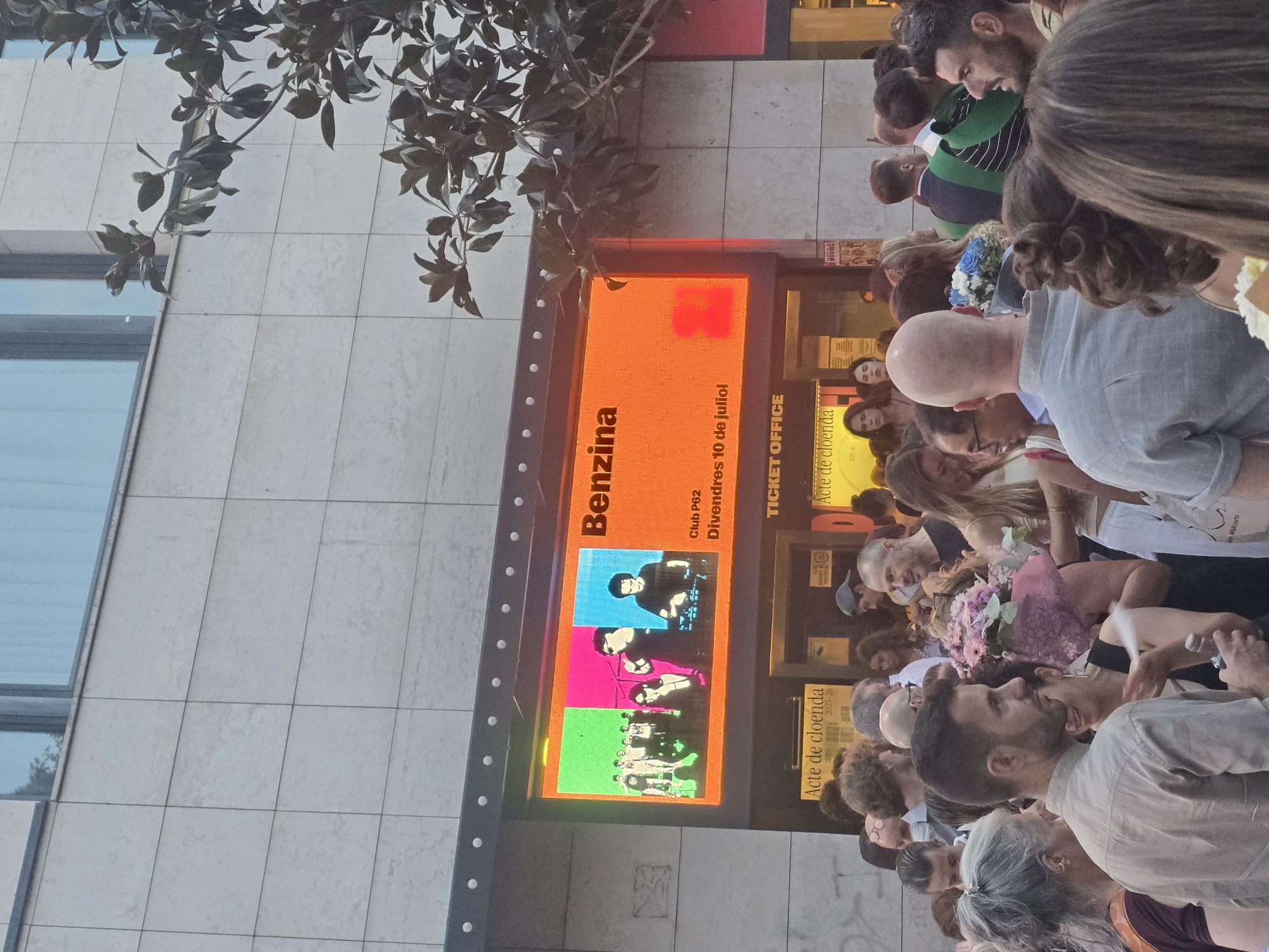 This media file captures a crowd of people gathered outside a building, likely for an event. The scene is oriented vertically, with the top of the image showing a portion of a building with a tiled facade and a window reflecting a tree. Below this, a brightly lit orange digital display is prominently featured, advertising "Benzina Club P62" and stating "Divendres 10 de juliol." The display also shows a series of colorful images, possibly related to the event. To the right of the main display, there are further posters and signs, one of which reads "TICKET OFFICE" and another "Acte de cloenda 2025-26."
The crowd consists of a diverse group of individuals of various ages, with many looking towards the entrance or the display. Some people are holding flowers, suggesting a celebratory or farewell event. The expressions on their faces are varied, but a general sense of anticipation and engagement is evident. The lighting suggests it is daytime, possibly late afternoon or early evening, given the warmth of the orange display. The tiled building and the Spanish text on the signs point to a location in Spain, specifically Barcelona as indicated. The overall atmosphere appears to be one of a public gathering related to a specific event or closing ceremony.