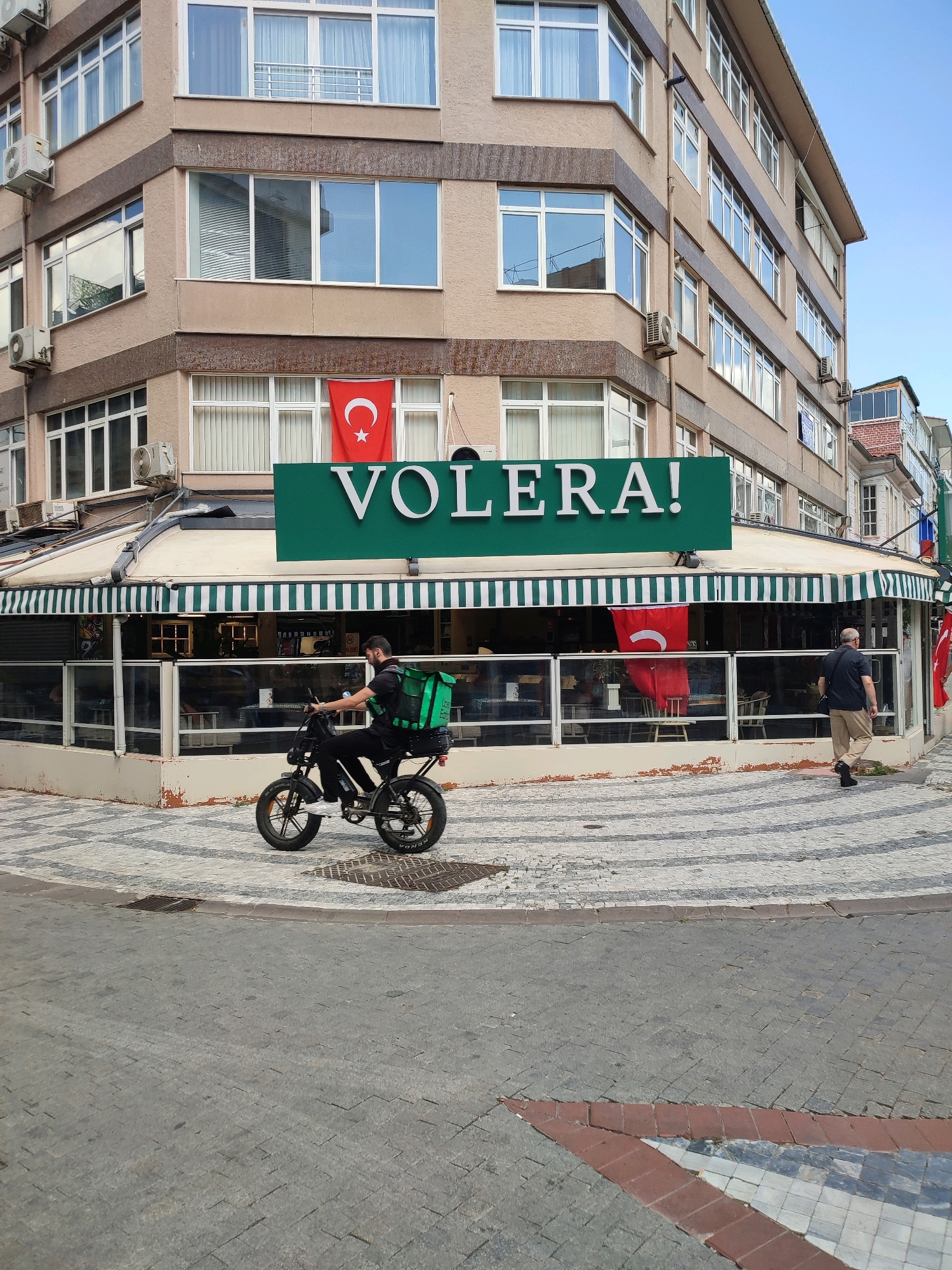 The image depicts a street scene in Kadıköy, Turkey, with a focus on a corner building housing a restaurant named "VOLERA!". A delivery person on a black electric motorcycle, equipped with a large green insulated bag on the back, is riding past the restaurant. The restaurant has a green awning with white stripes and large glass windows. Inside the restaurant, tables and chairs are visible. Two Turkish flags are displayed on the building's facade, one on the upper floor and another hanging near the restaurant's entrance. A man in a dark shirt and beige pants is walking away from the restaurant on the sidewalk. The street is paved with cobblestones and decorative patterned tiles. The building is multi-story, with numerous windows and air conditioning units visible on its exterior. The sky is clear and blue.