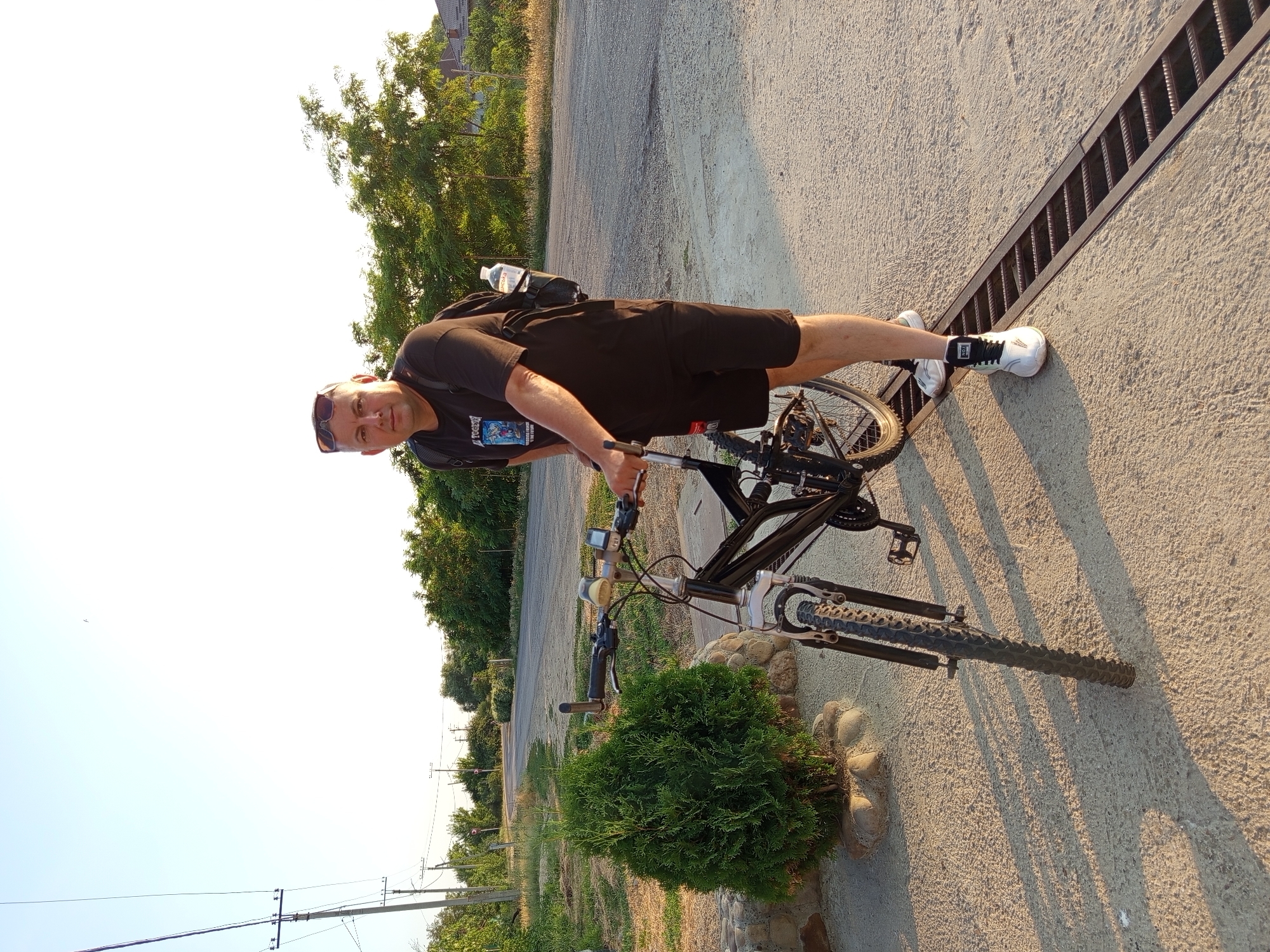 A man in a black t-shirt and black shorts stands with a black bicycle on a concrete surface next to a road. He is wearing sunglasses and a backpack with a water bottle attached. A small green bush is to his left, and trees and houses are visible in the background. The scene appears to be in Kitskany, Moldova, on a sunny day.