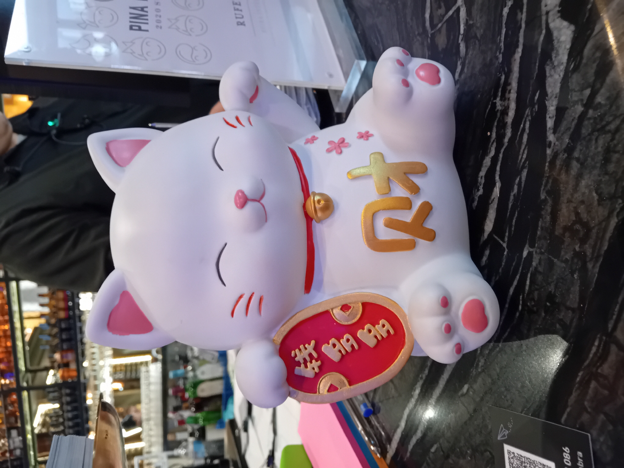 A white Maneki-neko (lucky cat) figurine lies on its back on a dark, veined marble or granite table. The cat has a white body, pink inner ears, and pink pads on its paws. Its eyes are closed with red markings, and it wears a red collar with a golden bell. Its right paw is raised, holding a red oval tablet with golden Chinese or Japanese characters; one clearly visible character is "吉" (meaning good fortune or luck), while others on the tablet are less distinct. On the cat's belly, large golden characters spell "開運" (meaning good luck or fortune), surrounded by small pink, cherry blossom-like designs.
The setting appears to be an indoor commercial establishment, possibly a restaurant, bar, or shop, indicated by blurred shelves with bottles and other items in the background. A person in a dark shirt is partially visible behind the cat, with a green light emanating from a device near them. An acrylic display stand or menu holder is present in the upper left background, featuring the text "PINA" and "2020 S", along with cartoon-style faces and the partial word "RUFE". In the lower right, a QR code and fragmented text including "986" and "bra" are visible on a piece of paper or card. The time of day is not discernible, but the scene is brightly lit by artificial indoor lighting. No specific activity is depicted beyond the static arrangement of objects.