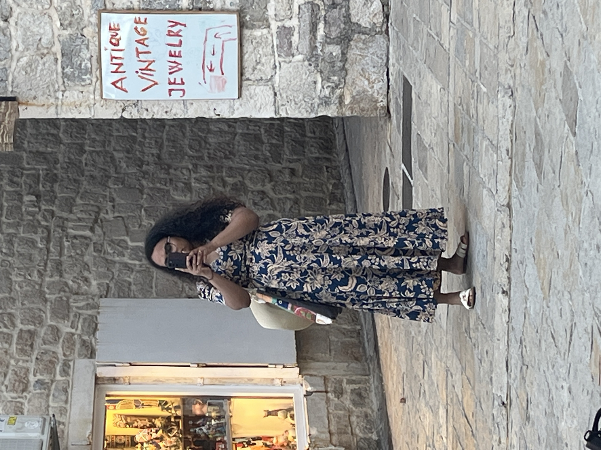 A woman with dark, curly hair and glasses is standing on a stone-paved street in Budva, Montenegro. She is wearing a long, dark blue dress with a floral pattern and white sandals. She is holding a smartphone in her hands, appearing to take a photograph or record a video. A light-colored straw hat with a patterned strap is slung over her left shoulder.
To her left, there is a shop window displaying various items, possibly souvenirs or trinkets, illuminated by warm light. The shop's facade is made of stone.
To her right, a white sign with red lettering reads "ANTIQUE VINTAGE JEWELRY". Below the text, there is a simple red line drawing that appears to depict jewelry or a jewelry item. The sign is mounted on a rough stone wall. The environment suggests an older, possibly historical part of the city, characterized by the stone architecture and paving. The lighting is subdued, indicating it might be late afternoon or early evening.