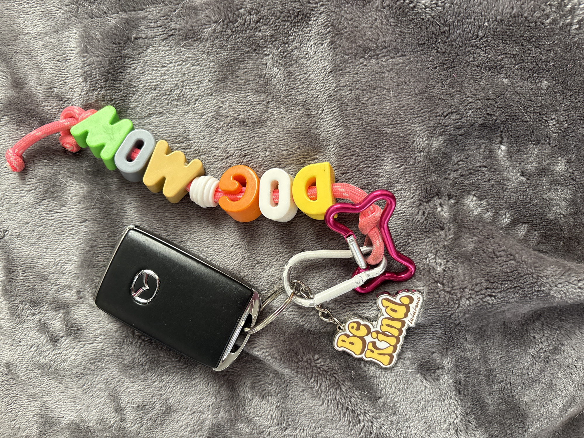 The image depicts a black Mazda car key fob attached to a keychain lying on a gray textured surface, possibly a blanket or soft fabric. The keychain features colorful letter beads spelling "MOM DOG" strung on a pink cord, interspersed with white spacer beads. A pink heart-shaped carabiner clip and a white carabiner are attached to the keychain, along with a "Be Kind" charm. The scene is not identifiable as being in Seal Beach, United States of America.