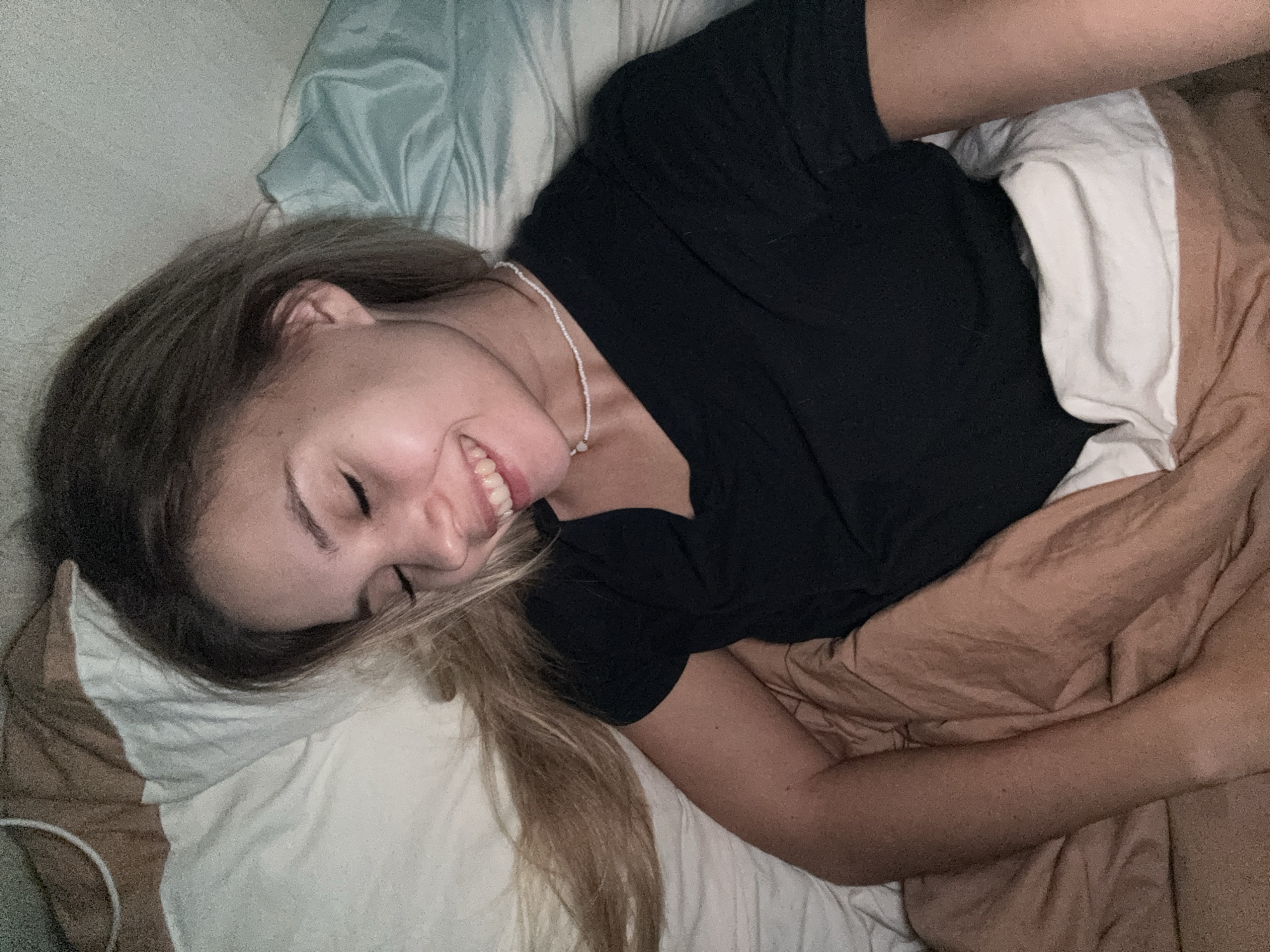 A young woman is depicted lying in bed, smiling broadly. She is positioned on her side with her head resting on a white pillow, and her long, light brown hair spreads across the pillow. She wears a black short-sleeved t-shirt and a thin white beaded necklace with a small pendant. Her eyes are partially closed, contributing to her relaxed and happy expression, highlighted by her wide, open-mouthed smile revealing her teeth.
She is covered by a light brown or tan duvet or blanket, with a white sheet visible underneath. Behind her head, part of a light blue patterned sheet or duvet cover can be seen. The setting appears to be a bedroom, characterized by the bedding and soft, somewhat dim lighting, possibly suggesting early morning or evening. The woman's joyful expression indicates a moment of happiness or contentment. No text is visible in the media file.