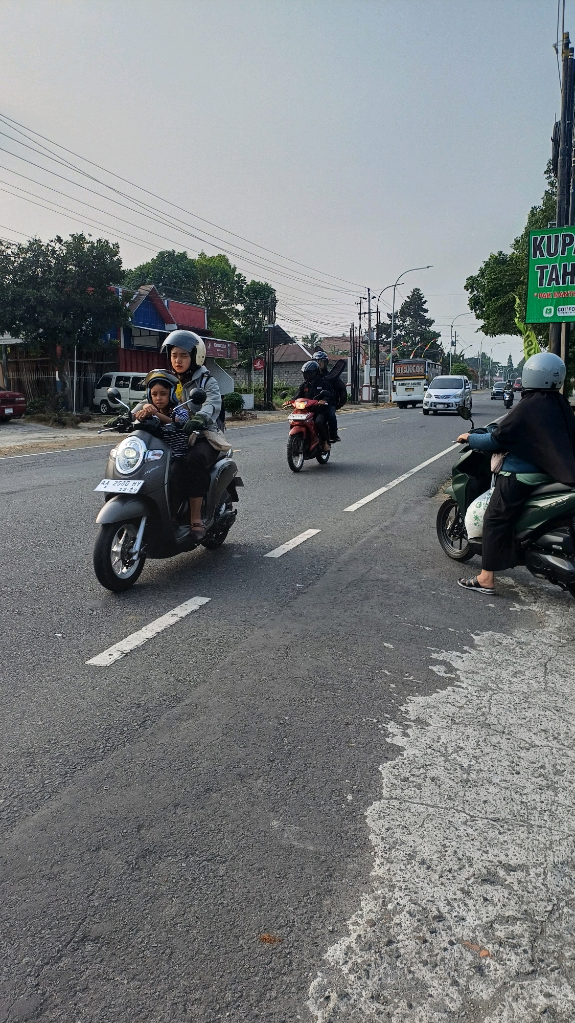 In the city of Badran, Indonesia, on a paved road under an overcast sky, two individuals ride a gray scooter in the foreground. The driver, wearing a white helmet and a gray jacket, is looking forward. A child, also wearing a helmet, sits in front of the driver. The scooter has a license plate with the characters "AA 2580 HY" and the date "12-25".
Behind them, on the same road, two other people are riding a red scooter. The driver, wearing a helmet, is looking forward. The passenger, also with a helmet, is looking to the side.
Further ahead, on the right side of the road, a person wearing a black hijab and a light blue shirt is riding a green scooter. They are looking to the left. In the background, several cars and other vehicles are visible on the road, along with buildings and trees lining the street. A green sign with white text is visible on the right side of the image. The road surface is asphalt with white lane markings.