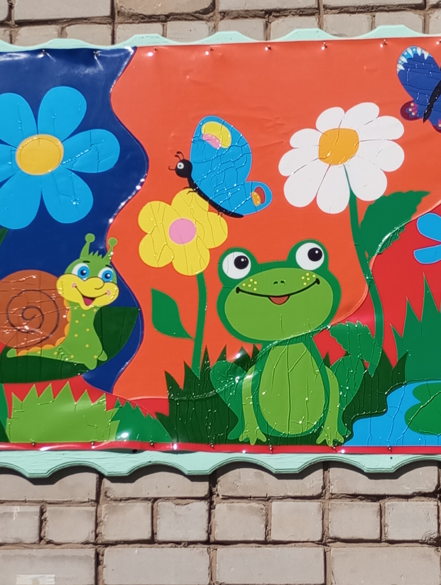 A close-up shows a colorful, cartoonish mural attached to a brick wall. The mural features a snail, flowers, butterflies, and a frog. The setting suggests the image was taken at a place where children play or learn. The mural is mounted with a border showing signs of wear. The brick wall has a pale grey color. There is no other context available to indicate where this may have been taken in Izhevsk, Russia. The environment is sunny.