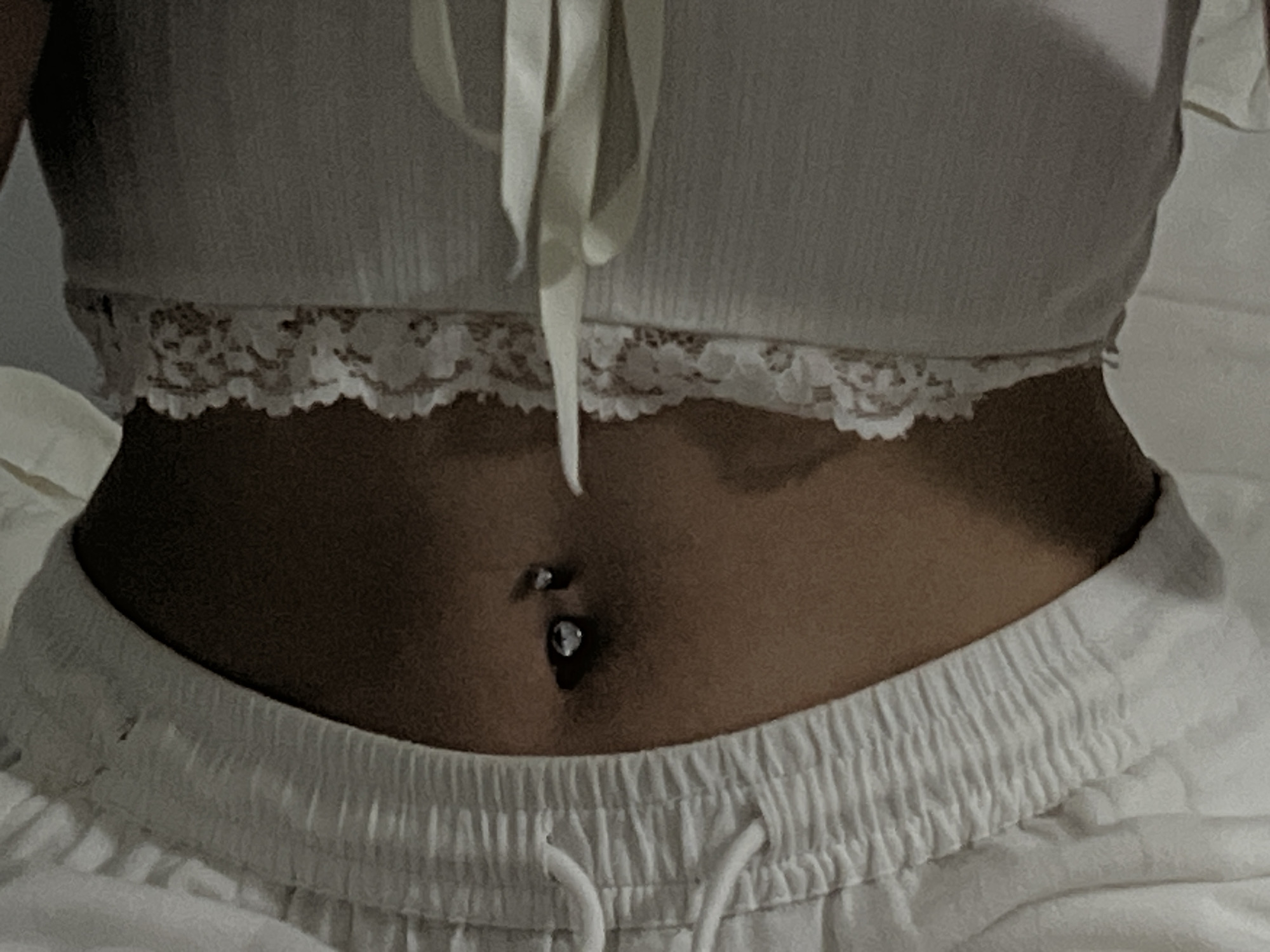The image shows a close-up of a person's midriff, with a navel piercing visible. The person is wearing a white lace-trimmed crop top and white drawstring pants. The background is out of focus and appears to be indoors, possibly a bedroom. There is no indication of the location in Ternopil, Ukraine.