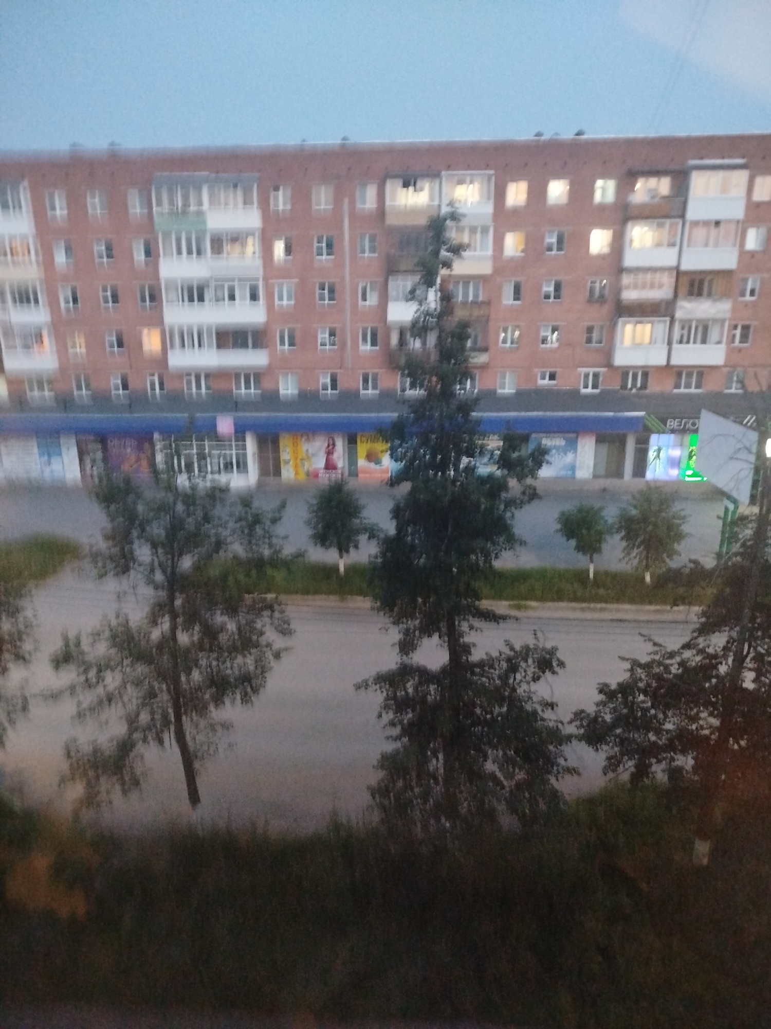 This media file depicts an exterior view of a multi-story residential building with commercial spaces on the ground floor, set in Votkinsk, Russia. The building is constructed of brick and features numerous windows and balconies. On the ground floor, several storefronts are visible, some displaying advertisements and signage. One sign, illuminated in green, appears to read "ВЕЛО" which translates to "BIKE" in Russian, suggesting a bicycle-related business.
The scene is captured during dusk, indicated by the soft, diffused light and the pale sky. The weather appears to be overcast or twilight, contributing to the subdued atmosphere. In the foreground and mid-ground, several trees are present, some with dense foliage and others appearing sparser. The trees are partially obscuring the view of the building and the street below. The overall setting appears to be a residential area with urban amenities. There are no people or specific activities clearly discernible in the image, giving a sense of a quiet, ordinary evening. The image evokes a calm and slightly melancholic mood due to the twilight lighting and the stillness of the scene.