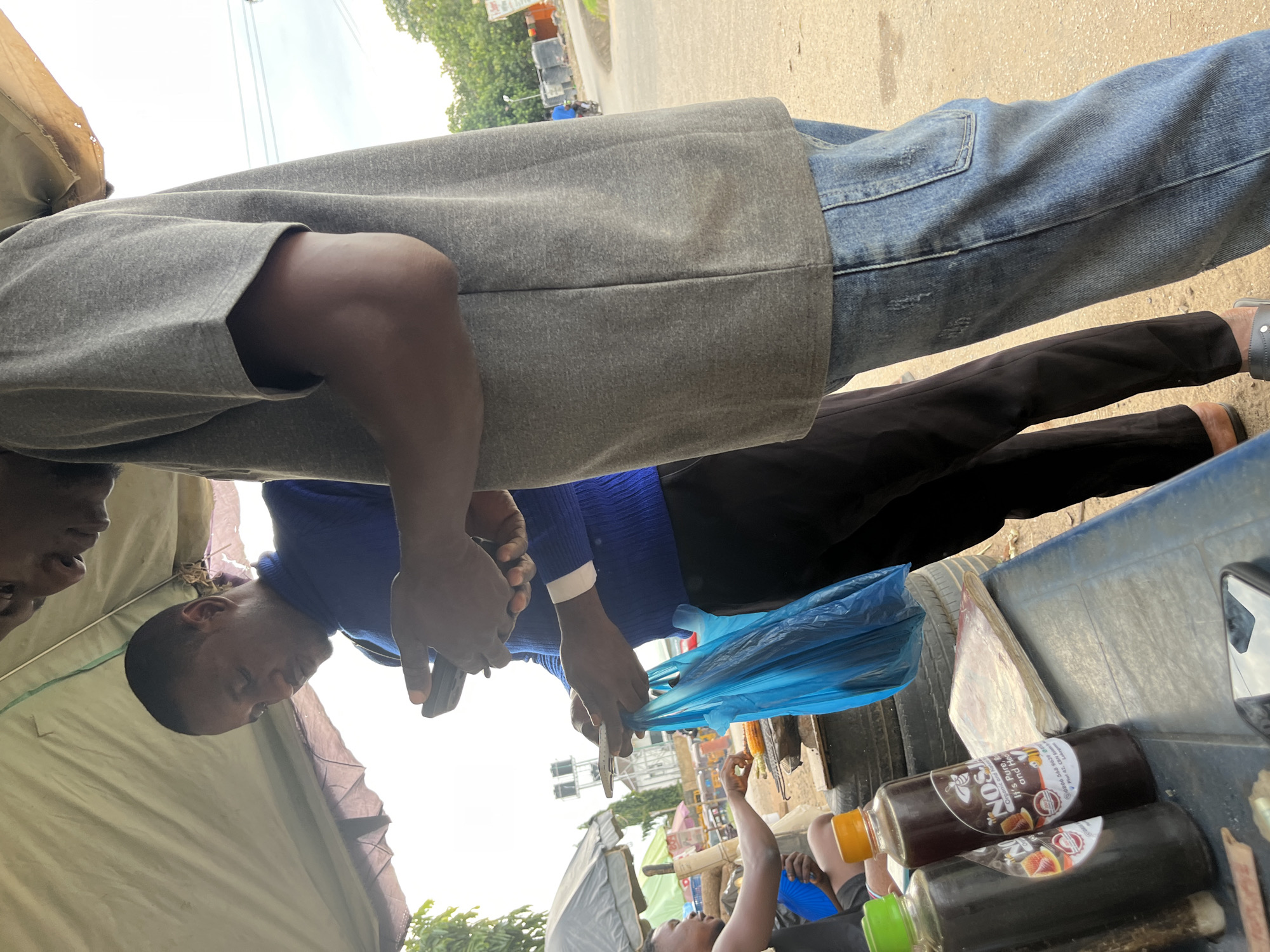 Two men are standing under a tent outdoors, appearing to be at a roadside stall in Abuja, Nigeria. The man on the left, wearing a blue sweater and black pants, is looking at his phone while holding a blue plastic bag in his left hand. He is positioned near a grill where another person, partially visible on the far left, is tending to food. The man on the right, wearing a grey t-shirt and blue jeans, is also looking down, presumably at his phone. On a table in the foreground, there are two bottles of what appears to be honey, along with other items. The environment suggests a typical street-side vendor setting with a mix of urban and somewhat rural elements visible in the background, including trees, buildings, and what might be vehicles. The lighting is overcast, indicating a cloudy day.