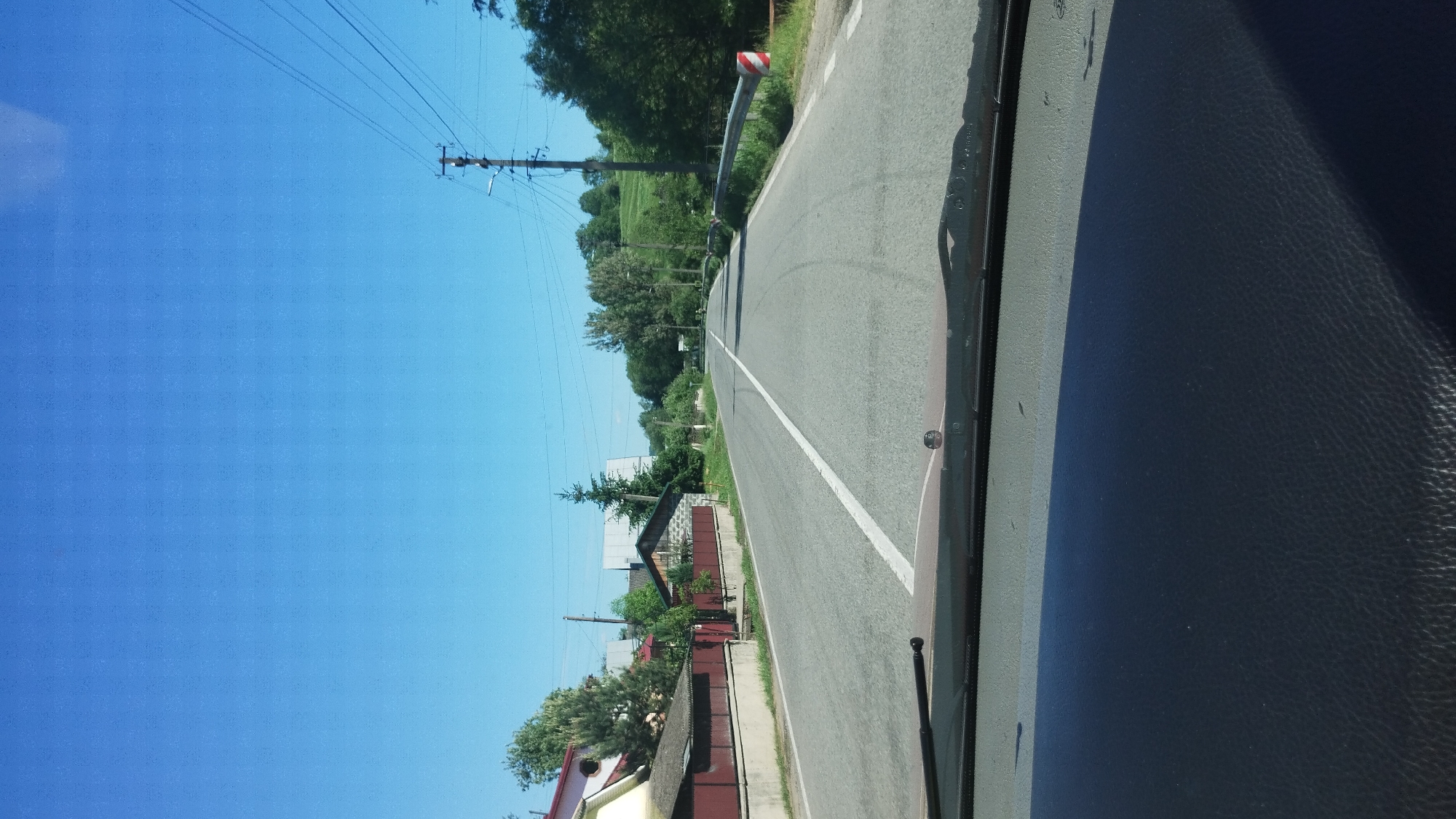 The image depicts a road in Deliatyn, Ukraine, from the perspective of someone inside a vehicle. The road is paved and has a white line marking the lane. On the right side of the road, there is a dark, textured surface, likely the dashboard of the vehicle. On the left side of the road, there are houses with red roofs and stone foundations, surrounded by greenery and trees. Power lines run along the left side of the road, supported by wooden poles. The sky is a clear, bright blue, indicating a sunny day. There is a sign on a curved pole near the side of the road with red and white stripes, possibly a warning sign. The overall scene suggests a rural or suburban setting during daytime. No people are visible in the image, and no text is discernable.