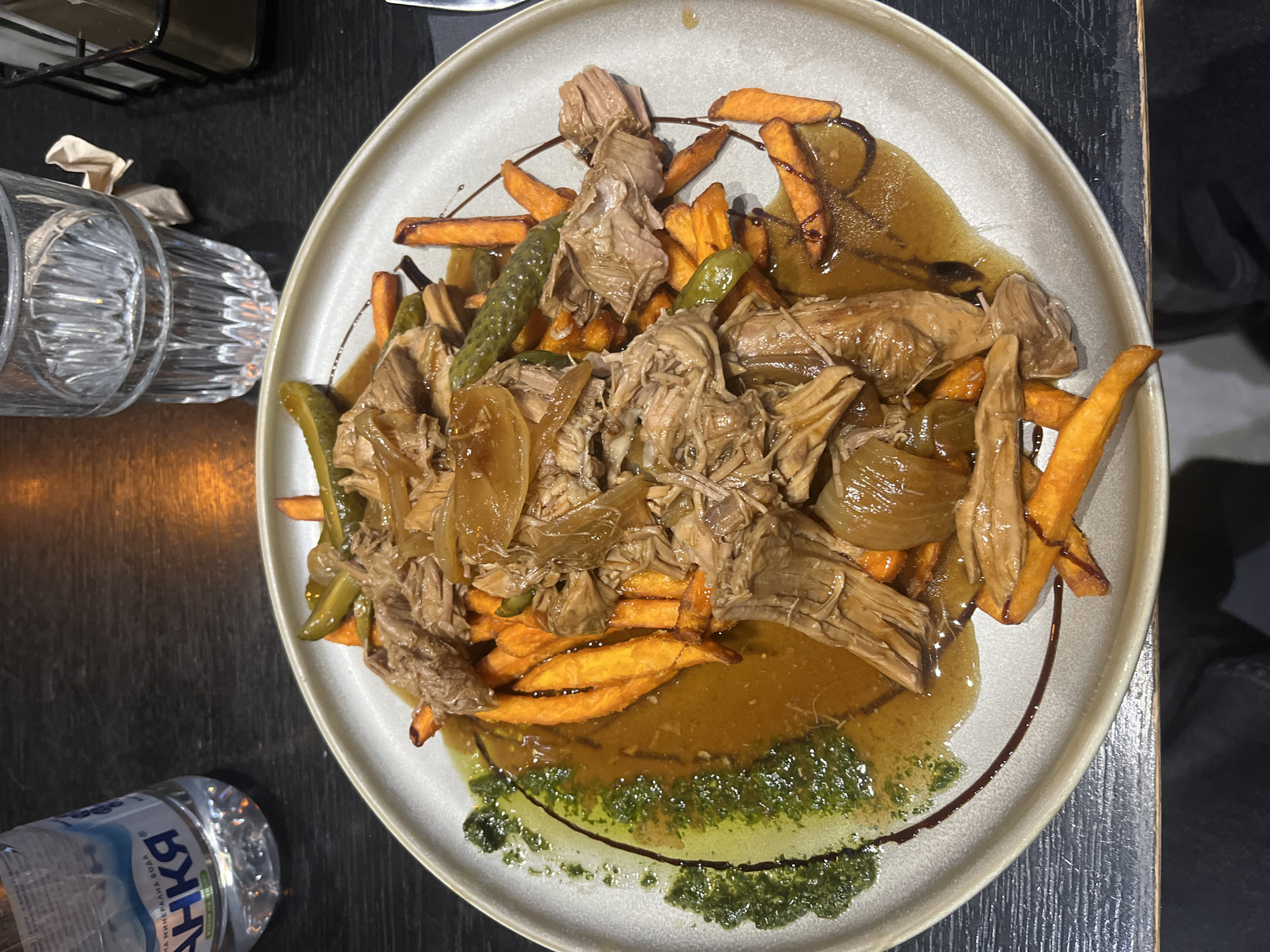The image shows a plate of food on a dark-colored table. The plate contains shredded meat with sauteed onions, sweet potato fries, and what appear to be pickled green beans. The food is dressed with two sauces, one is brown and gravy-like, the other a bright green pesto. The edge of the plate is light gray. A partially empty water bottle and an empty glass are on the left of the plate on the table. The photo appears to have been taken indoors, in a restaurant setting. The location in the city of Blagoevgrad, Bulgaria is not discernible from the information provided in the image.