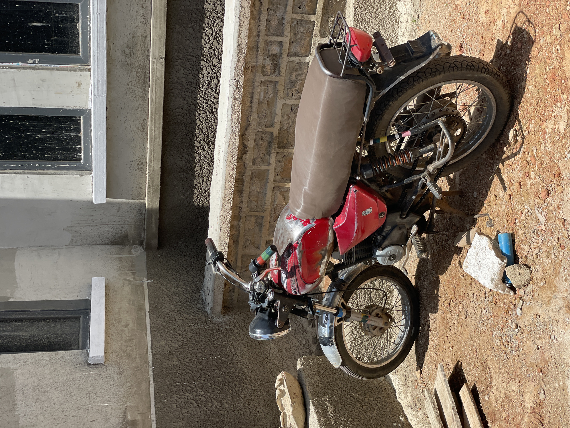A red and silver motorcycle is parked on a dirt lot in front of a partially constructed building. The motorcycle has a brown seat, a chrome headlight, and a rear cargo rack. The building has a rough, grey stucco exterior with two windows. The ground is covered in reddish-brown dirt and small stones, with a few pieces of wood and a blue pipe visible in the foreground. The scene appears to be in an outdoor, construction area. This location is Bar Gada, Nigeria.