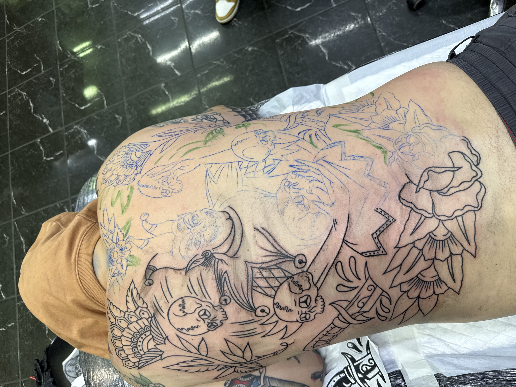 A high-angle shot shows a person lying on their side with a large tattoo design on their torso. The design includes floral motifs, skulls, and other graphic elements. Some parts of the tattoo are outlined in black ink, while other sections are sketched in blue and green. The person is wearing dark shorts and a tan-colored shirt, part of which is visible. The scene appears to be a tattoo studio, as indicated by the white paper covering the surface on which the person is lying, and the black tiled floor. There is no indication in the photo it was taken in Moscow, Russia.