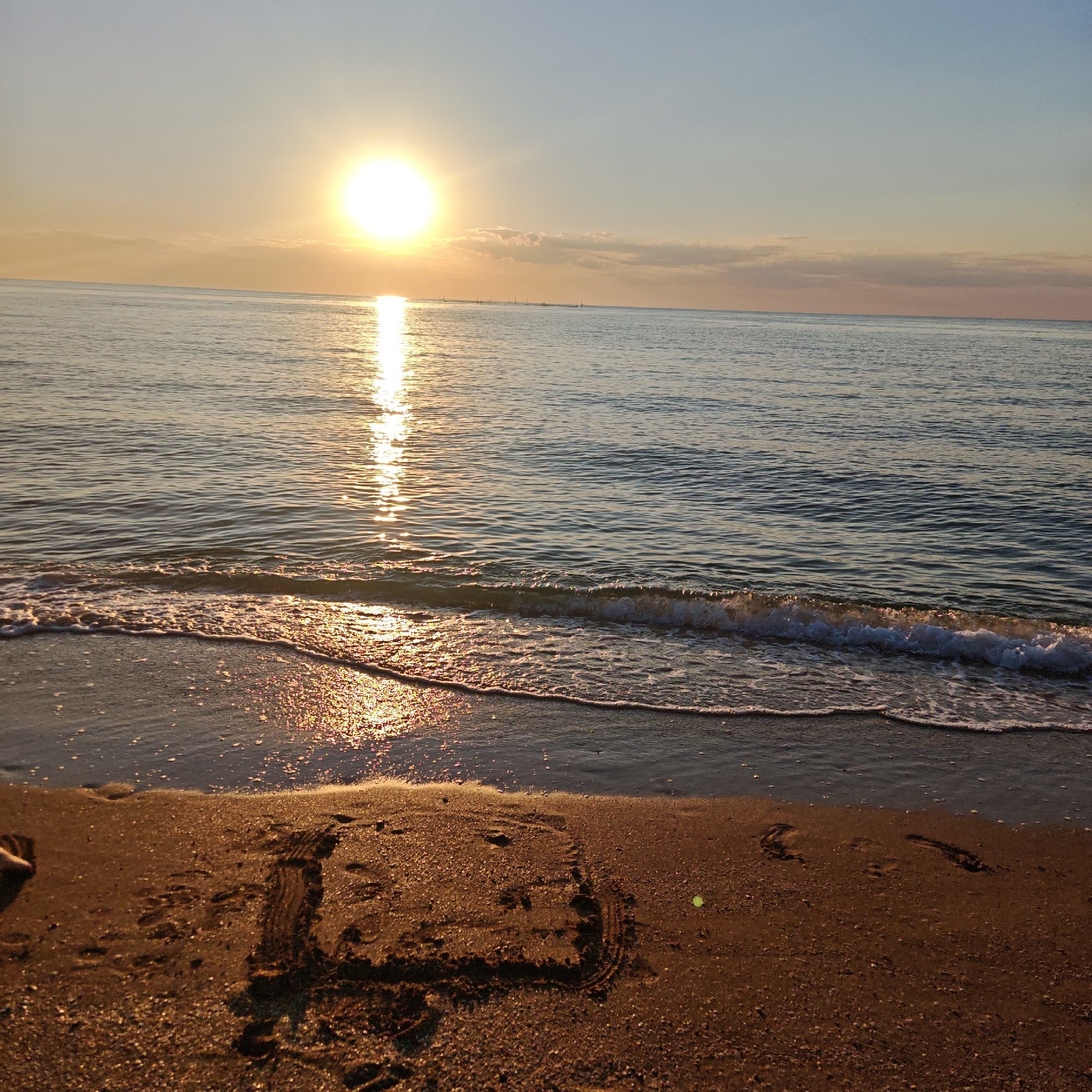 In a serene coastal scene, the sun hangs low in the sky, casting a golden glow across the calm ocean and wet sand. The sun's reflection shimmers on the water's surface, creating a path of light. Gentle waves lap at the shore, leaving a thin line of foam and glistening sand. On the beach, a child's drawing is etched into the wet sand, depicting a simple face with two eyes and a smile. Footprints are scattered nearby, hinting at previous activity. The sky transitions from a pale blue at the top to a warm, orange hue near the horizon, with soft clouds adding texture. The overall atmosphere is tranquil and peaceful, suggesting a quiet moment at sunrise or sunset. The location is unknown.