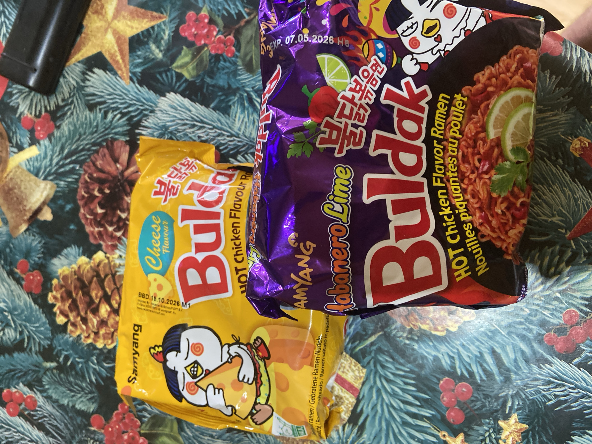 The image shows two packets of instant noodles on a Christmas-themed tablecloth. On the left is a yellow packet labeled "Samyang Cheese Flavour Buldak Hot Chicken Flavour Ramen". The packet features a cartoon chicken character holding a piece of cheese. The expiration date "BBD.19.10.2026 M1" is printed on the packet. To the right is a purple packet labeled "AMYANG Habanero Lime Buldak Hot Chicken Flavor Ramen Nouilles piquantes au poulet". The expiration date "EXP. 07.05.2026 H6" is printed on the packet. The background consists of a tablecloth with a pattern of pine branches, red berries, pine cones, and gold stars. The location, city of Sofia, Bulgaria, is not indicated in the photo or its crops.