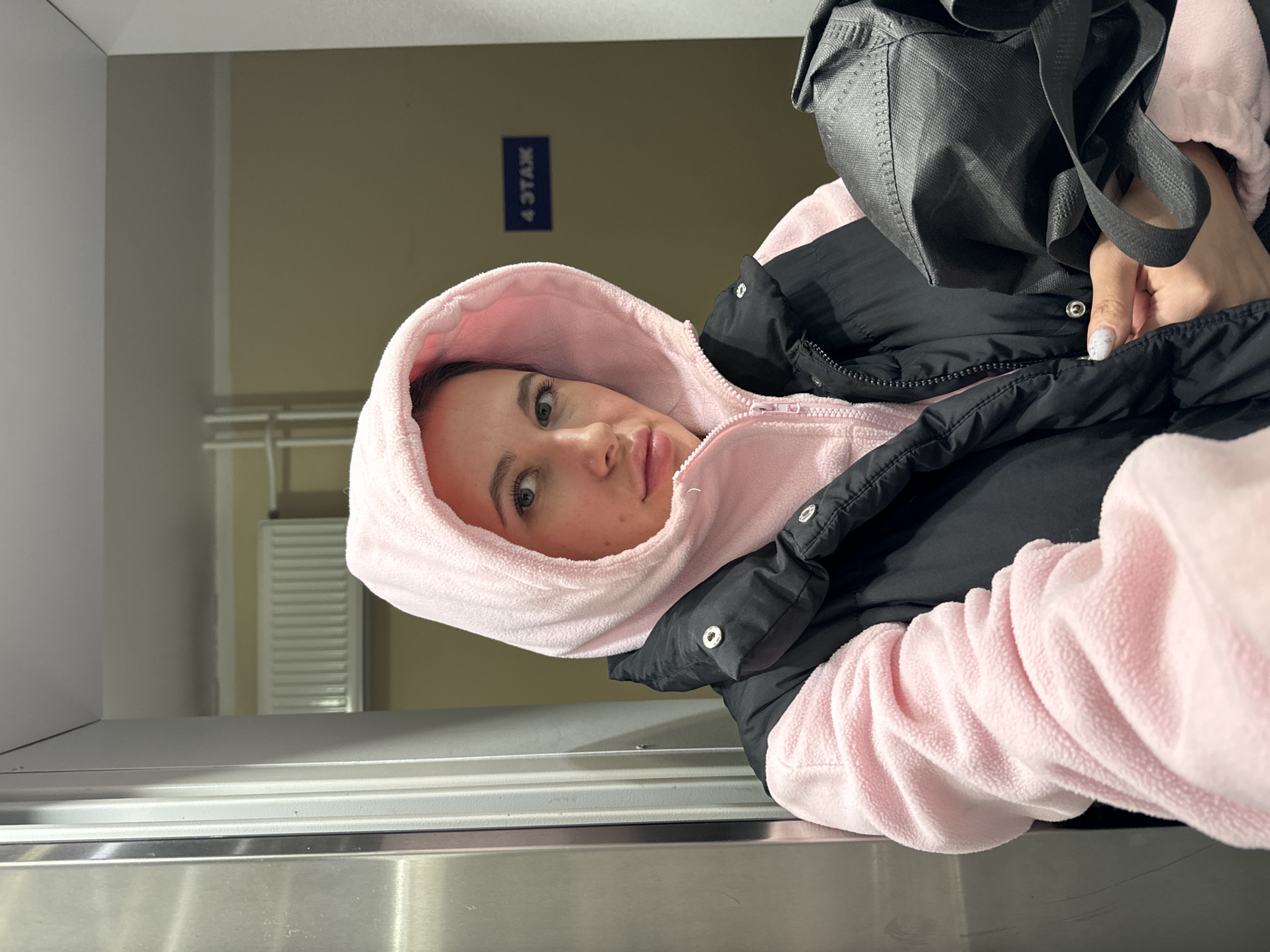 A single female individual is depicted in an indoor environment, likely within an elevator car or adjacent to one. The subject, a woman with dark hair and light eyes, wears a pink fleece hooded sweatshirt with the hood pulled up, layered under a black quilted vest. Her left hand, with manicured nails, holds a black, textured, possibly non-woven fabric bag. To the left, a metallic panel with vertical seams is visible. The background consists of a plain light-brown wall with a white ceiling and trim. A white radiator is mounted on the wall. A blue rectangular sign with white Cyrillic text reading "4 ЭТАЖ" (4th FLOOR) is affixed to the wall on the right. The scene is located in a building on the 4th floor in Moscow, Russia.