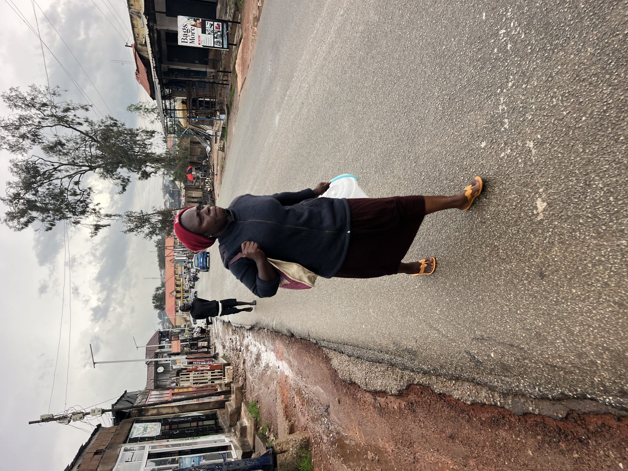 The image depicts a street scene with several people and buildings. A woman in the foreground is walking on the asphalt road, carrying a white plastic bag and a light-colored bag over her shoulder. She is wearing a dark blue jacket, a dark skirt, and an orange sandal on her right foot. She has a red head covering. To her left, another person, a man, is walking on the same road, wearing a black shirt and black shorts.
The setting appears to be an urban environment with shops lining the street. Some of the shops have signs with text, including "Bags More" and "Coca-Cola". There are also trees, power lines, and utility poles visible. The road is wet in some areas, suggesting recent rain. The sky is overcast with clouds, indicating a cloudy or possibly rainy day. The overall atmosphere is that of a typical street scene, possibly in a market area or a commercial district. The image is oriented vertically.