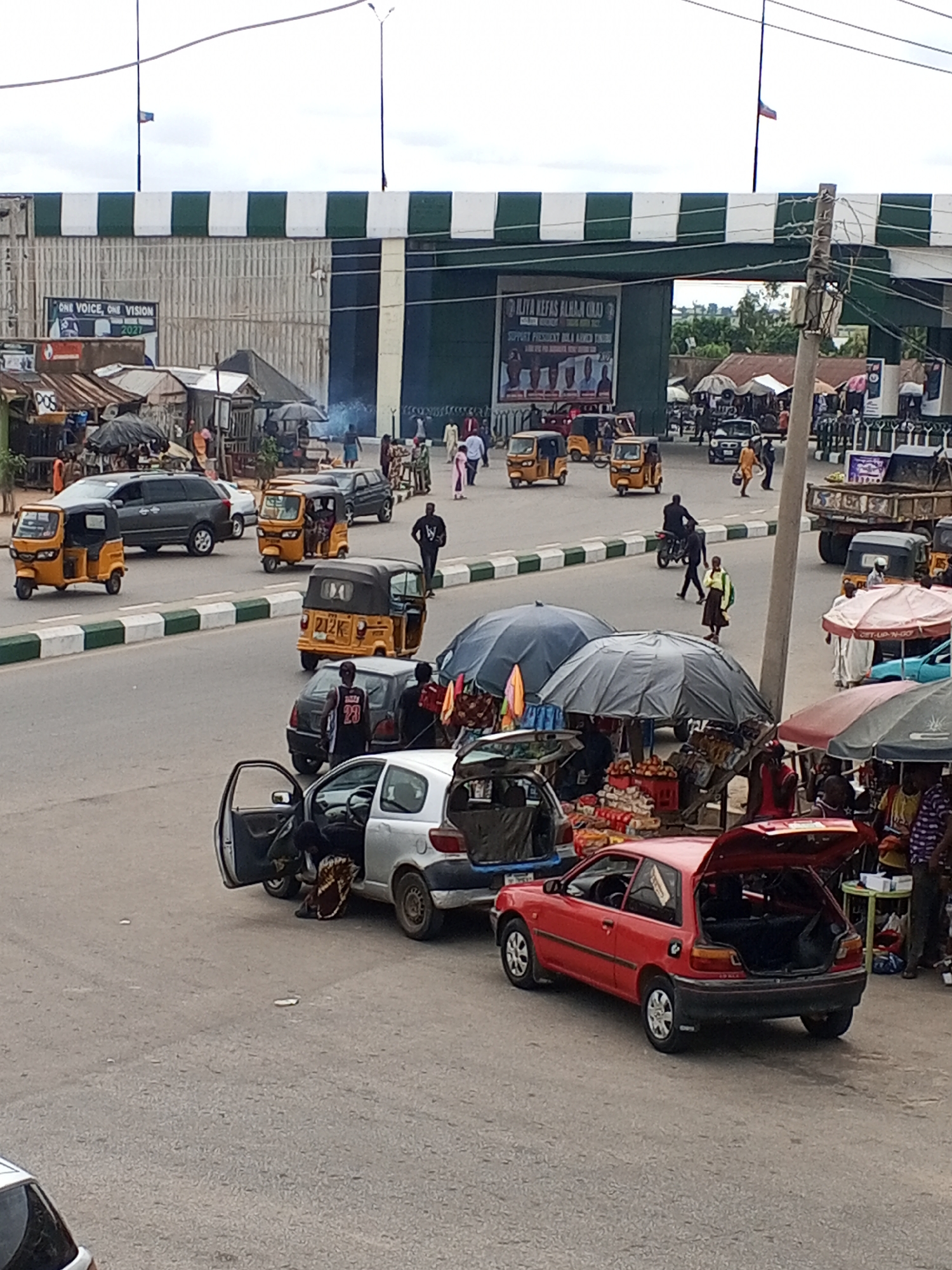The image depicts a street scene in Jalingo, Nigeria, characterized by a bustling market and heavy traffic. In the foreground, a red Toyota Starlet hatchback with its trunk open is parked, with its back passenger door also ajar. To its left, a silver Toyota Yaris is parked with its driver's side door open, and a person is partially visible inside.
The street is lined with market stalls covered by umbrellas, displaying goods such as fruits and packaged items. Several tricycles, commonly known as 'keke napeps', are navigating the road, along with a dark grey van and a light grey van in the middle ground. Further back, a large truck with a raised flatbed is visible, alongside more market stalls and a large archway adorned with banners. Pedestrians are seen walking on the sidewalks and crossing the street, some carrying items. The environment appears to be a busy commercial area with an overcast sky. The overall impression is one of everyday life in a Nigerian city, with commerce and transportation in full swing.