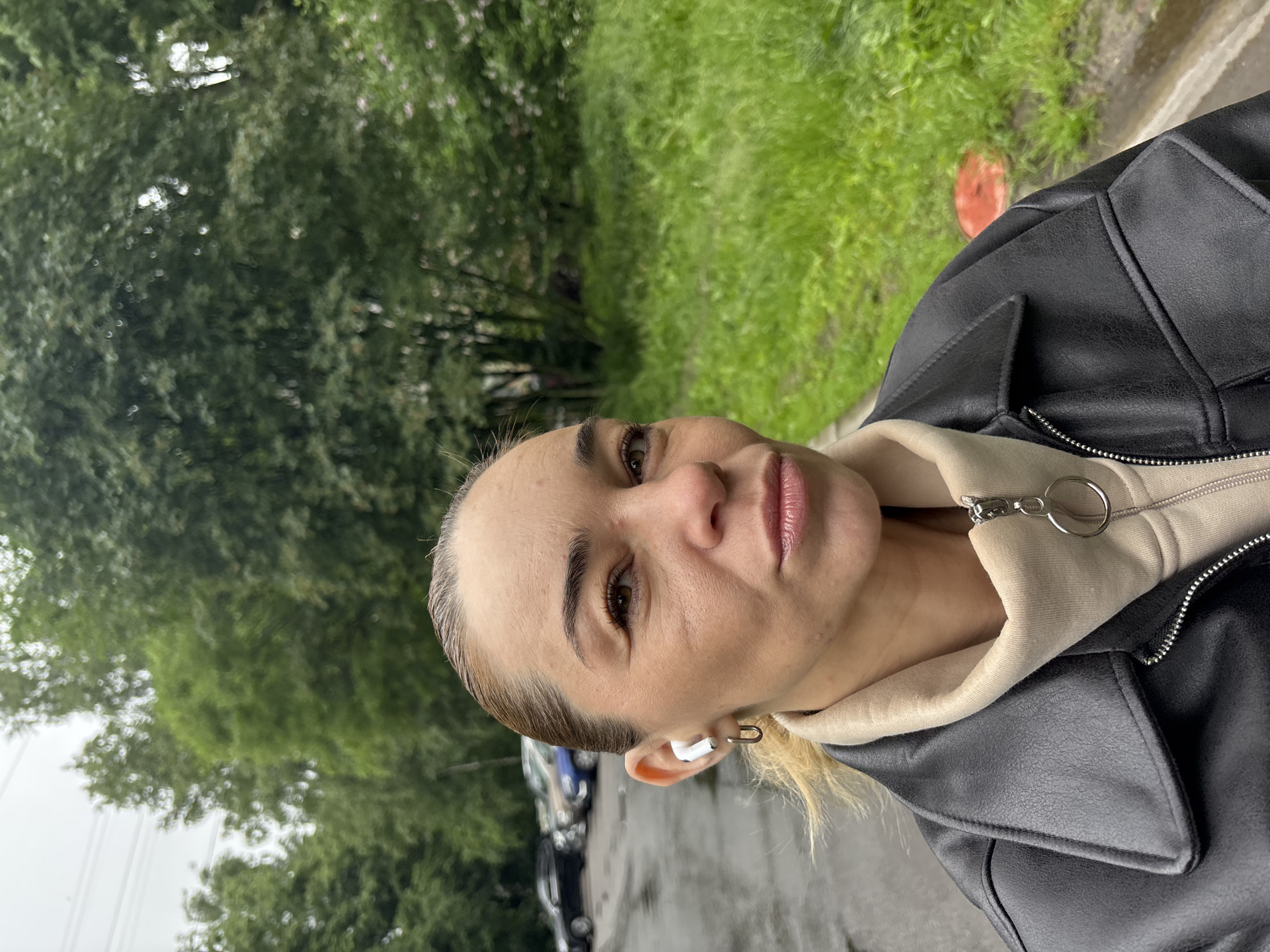 The media file is a portrait of a woman outdoors in Vsevolozhsk, Russia. The woman is wearing a black jacket over a beige hoodie and white earbuds. She has blonde hair and appears to be in her late 30s or early 40s. Her gaze is directed slightly to the left of the camera, and she has a neutral expression.
The background consists of lush green trees and grass, suggesting a park or a residential area with significant greenery. The ground appears to be wet and reflective, possibly indicating recent rain or a damp environment. Some cars are visible in the background, blurred and out of focus, adding context to the urban or suburban setting. A bright orange object, possibly a piece of litter or a fallen leaf, is visible on the grass.
The lighting suggests it is daytime, likely overcast or late in the day given the soft light. The image is taken from a slightly low angle, looking up at the woman. The overall mood is casual and relaxed, possibly capturing a moment during a walk or outdoor activity. There is no visible text in the image.