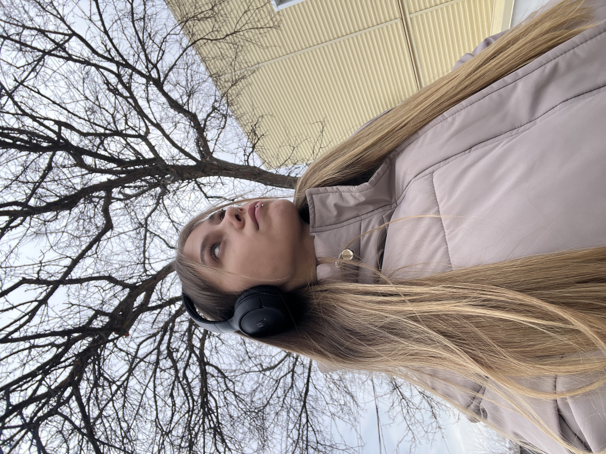 A young woman stands outdoors in what appears to be the city of Фоки, Russia. The woman has blonde hair and is wearing a beige-colored puffer jacket and black headphones. She is looking up, and her face is partially visible. She has lip piercings. Behind her is a tree with bare branches reaching towards the sky, suggesting it might be winter or early spring. A building with a pale yellow facade is also visible. The image is taken from a low angle.