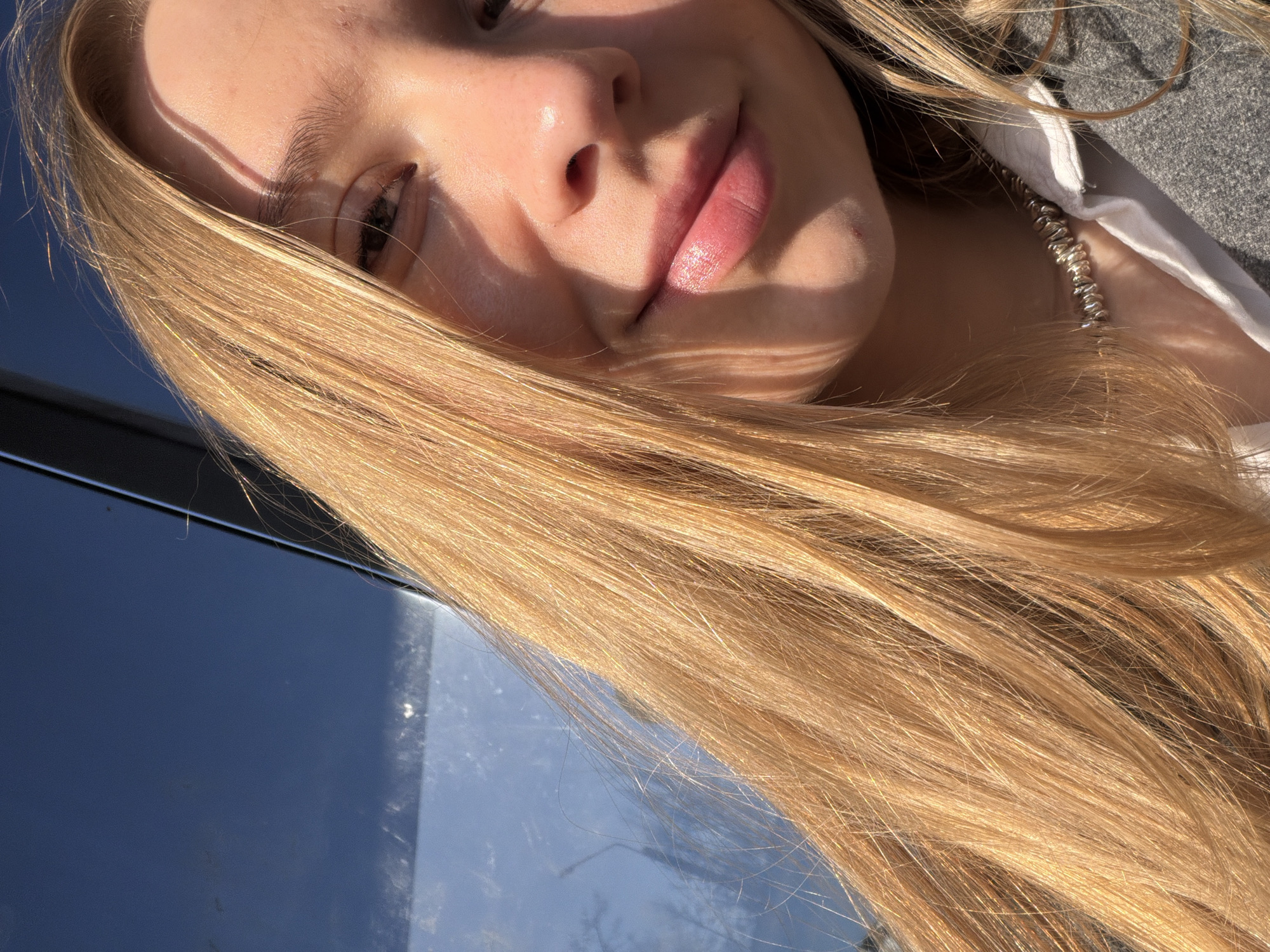 Close-up, tilted perspective of a fair-skinned person with long blonde hair, focusing on their face. The person is wearing a silver necklace, a white collared shirt, and a gray jacket. The background is a blue, reflective surface, possibly a window or a glossy surface, which reflects an indistinct outdoor scene. The image is illuminated by sunlight, creating sharp shadows on the person's face. It's impossible to confirm that the location is Chaikovsky, Russia. The image lacks context clues and landmarks. The provided information is based solely on visual data.