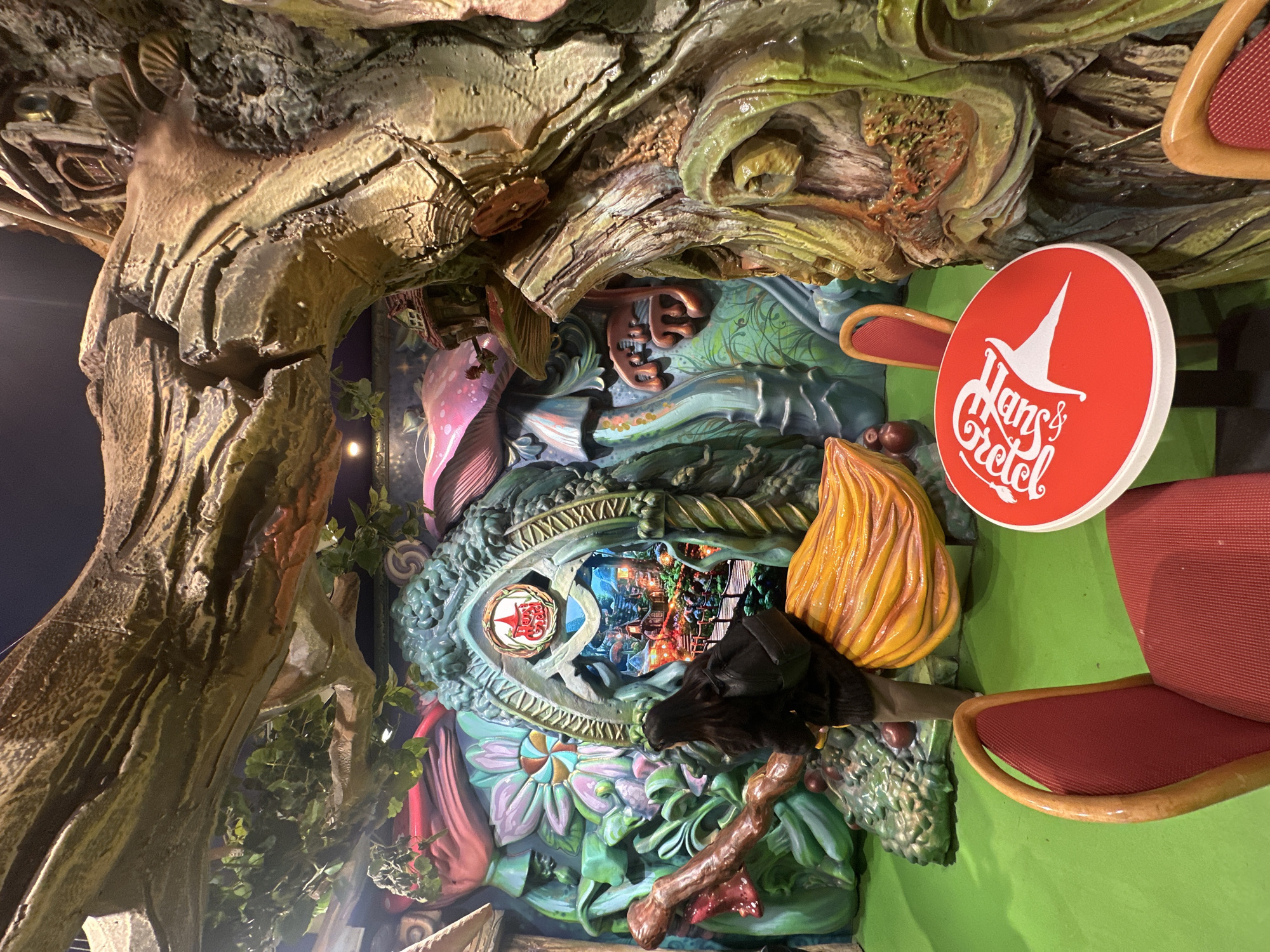 The image displays an indoor themed environment, identified by prominent branding as "Happy Land" in Plovdiv, Bulgaria. The scene is dominated by fantastical, oversized sculpted elements designed to resemble a vibrant, non-realistic forest setting.
A single individual, an adult female with long dark hair, is visible from behind, positioned center-left. She is wearing a dark jacket, dark pants, and a dark backpack. The individual is seated on a large, textured yellow sculpted object resembling a broomstick, facing a central, intricately sculpted archway.
The archway, featuring detailed organic shapes and a small structure resembling a house at its apex, frames an illuminated diorama depicting a colorful, fantastical village scene with multiple small buildings and glowing elements. A red circular sign displaying the "Happy Land" logo with a white conical hat graphic is integrated above the archway's opening.
The surrounding walls and ceiling are covered with large, sculpted elements including textured tree trunks and branches, some adorned with artificial green foliage. Oversized, vividly colored sculpted mushrooms (pink, blue, red) and various fantastical plant forms constitute the background. A substantial green sculpted mound containing multiple red spherical objects is situated near the broomstick. The floor is a uniform bright green.
In the foreground, a small round table with a red top prominently features a white "Happy Land" logo. Two red chairs with light brown wooden frames are arranged around this table.