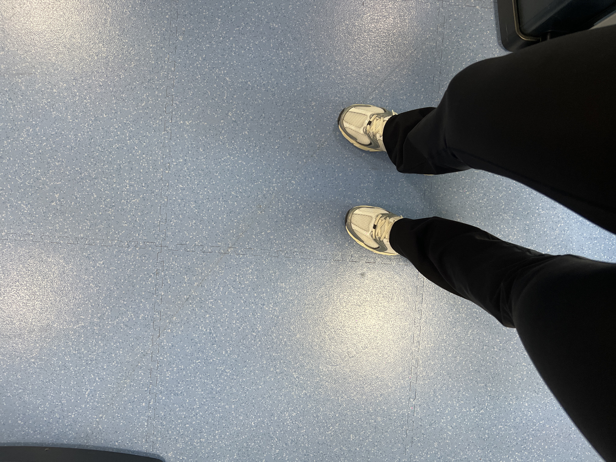 The image presents a first-person view looking down at a pair of feet clad in white sneakers and black trousers. The feet are positioned on a light blue floor comprised of interlocking, puzzle-piece-like tiles with a speckled pattern. The location is indeterminate, as the scene contains no clues that would definitively link it to Yerevan, Armenia. There are no people, actions, or interactions visible in the image. The environment consists solely of the tiled floor.