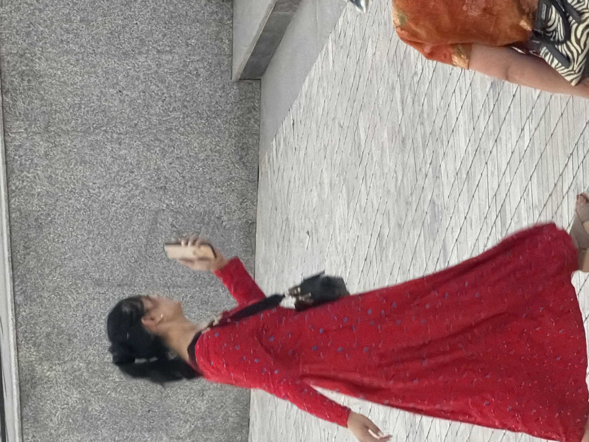 In an outdoor urban setting, a person wearing a red dress with blue polka dots walks to the left, holding a mobile phone in their right hand. The person's hair is pulled back in a ponytail, and they are wearing a small earring in their visible ear. A black crossbody bag is slung over their left shoulder. The ground is paved with light-colored rectangular tiles arranged in a diagonal pattern. Behind the person is a grey stone wall with a coarse texture. To the right of the frame, the shoulder and upper back of another person wearing an orange shirt with a floral pattern are visible. This person also has a bag with a black and white zebra print strap. The overall lighting suggests it is daytime.